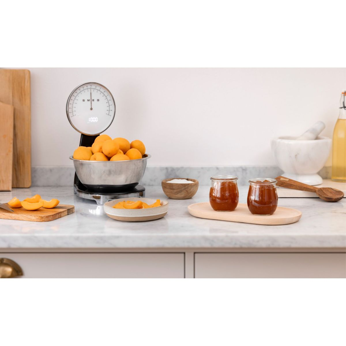 Blat kuchenny z wagą, morelami, słoikami dżemu i drewnianą łyżką. Scena jest jasna.