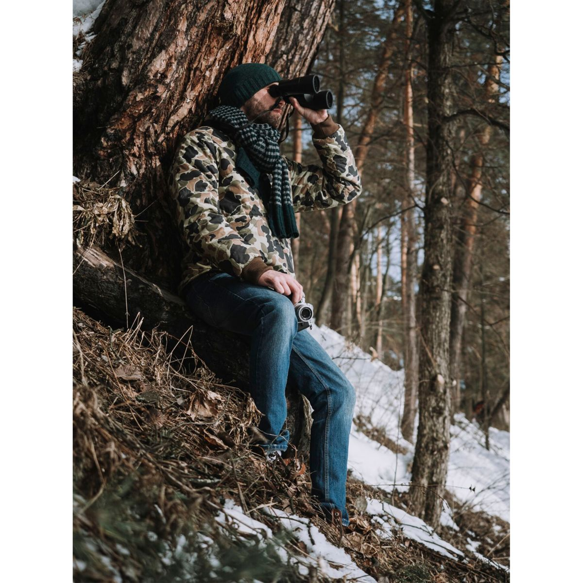 Mężczyzna w lesie z lornetką, w czapce i szaliku.