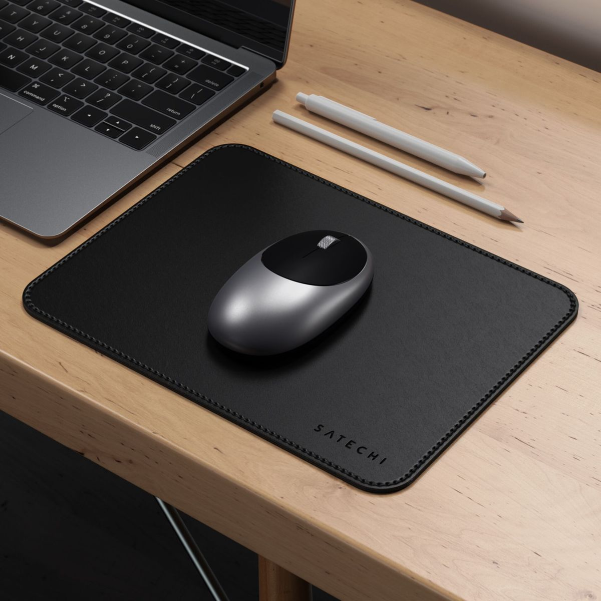 Ein schwarzes Mauspad mit einer silbernen Maus, einem Laptop und Stiften auf einem Holztisch.