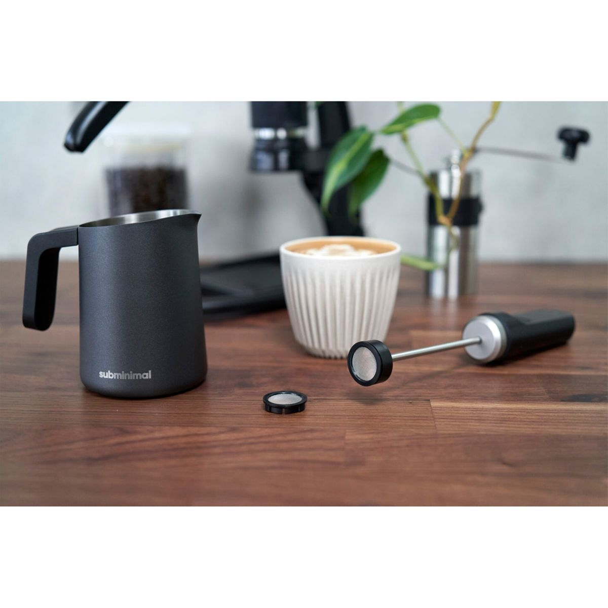 Eine Kaffeezubereitung mit schwarzem Krug, weißer Tasse, Milchaufschäumer und Mühle auf einem Holztisch.
