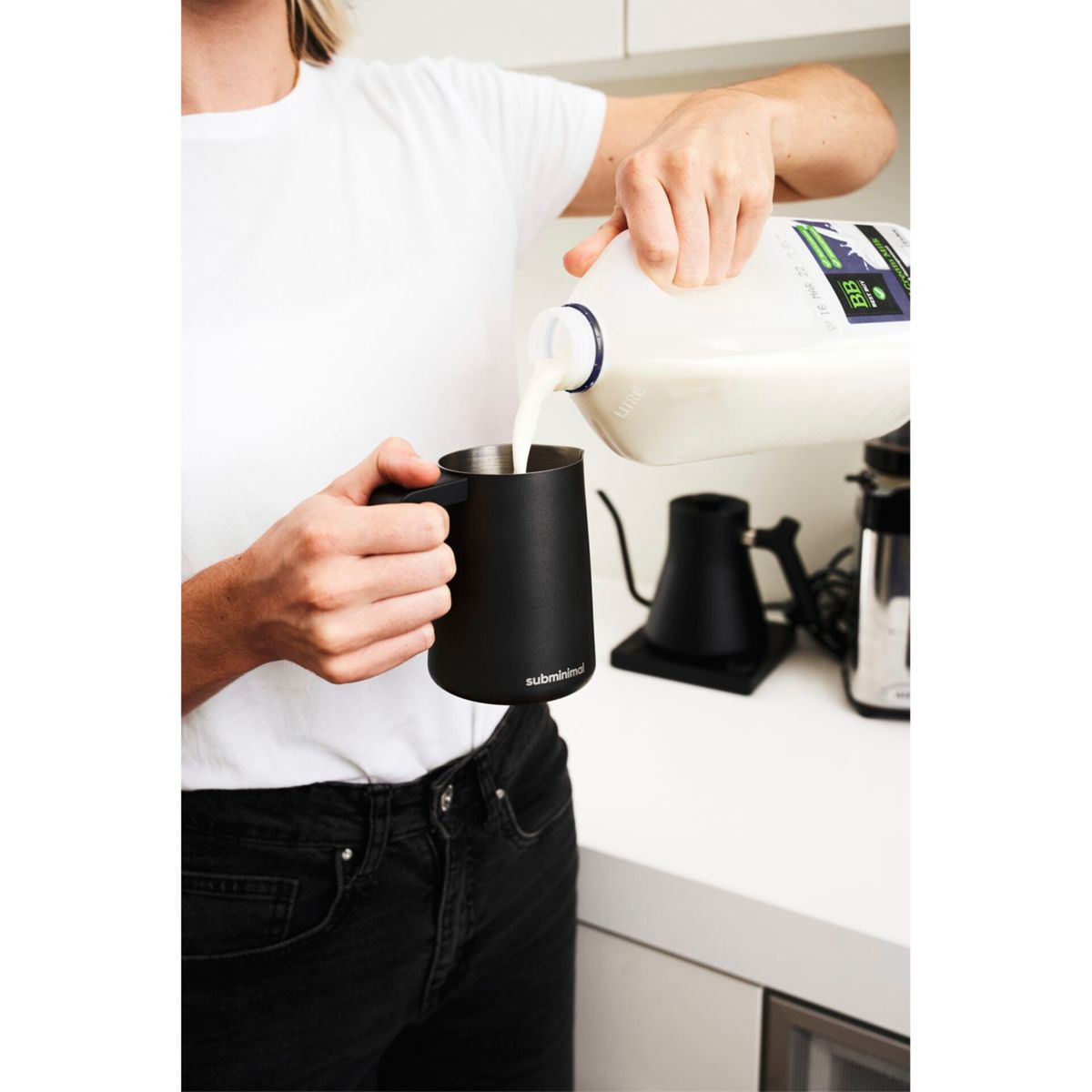 Eine Person gießt Milch in eine schwarze Tasse. Der Hintergrund zeigt eine weiße Theke und einen schwarzen Wasserkocher.