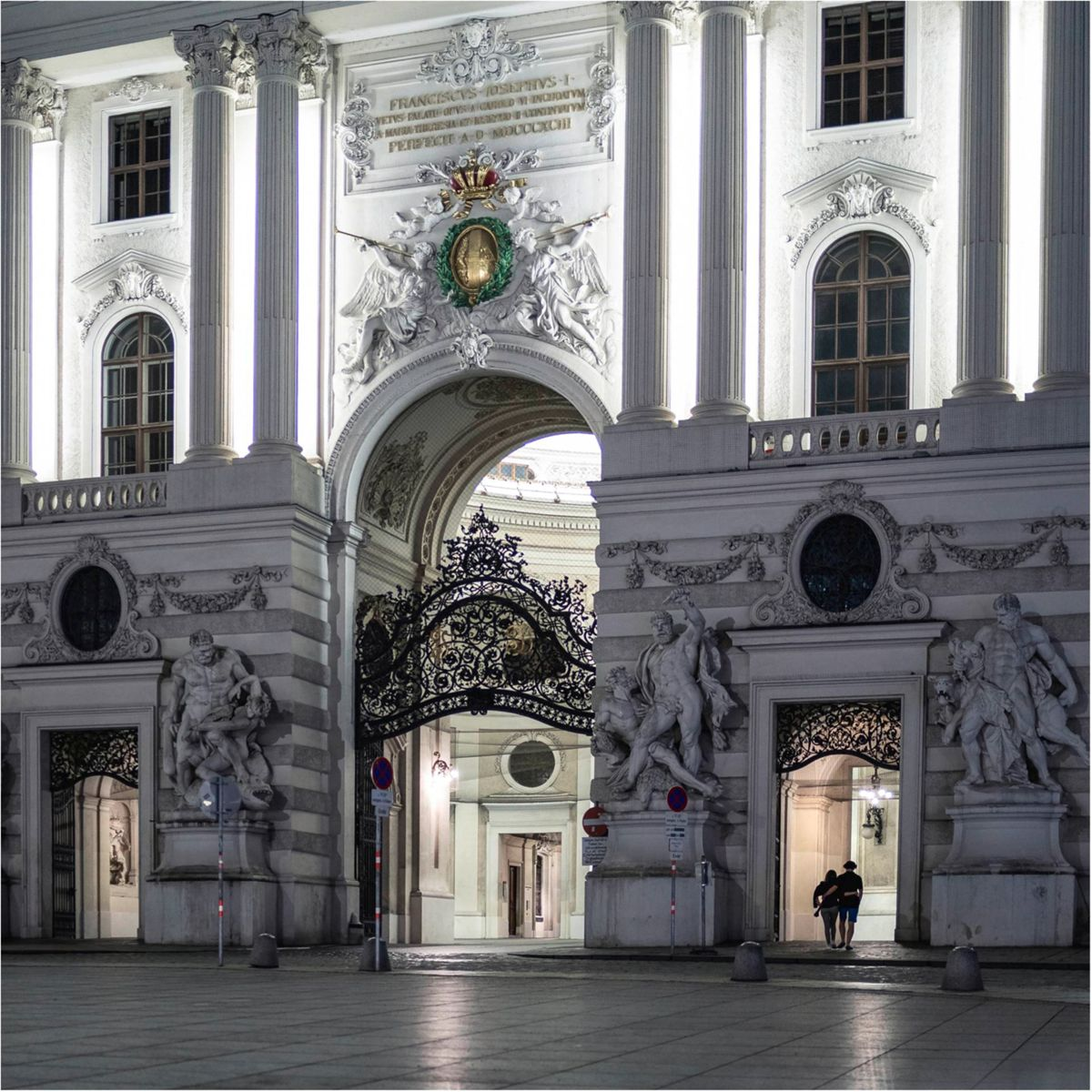 Nocne ujęcie wejścia do budynku z bogatą architekturą, posągami i bramą.