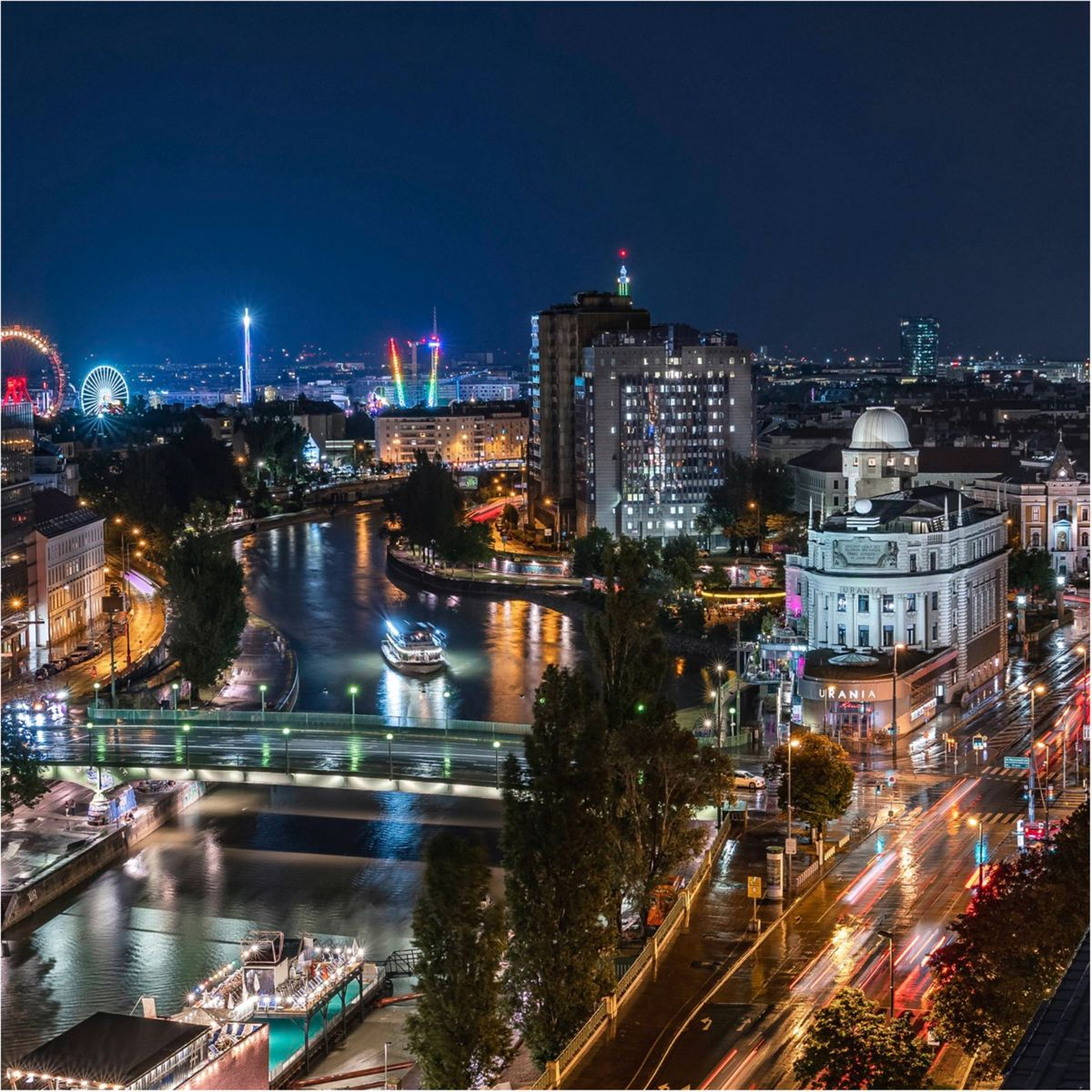 Nocny widok na miasto z diabelskim młynem i budynkami, rzeką z łodzią i światłami miasta.