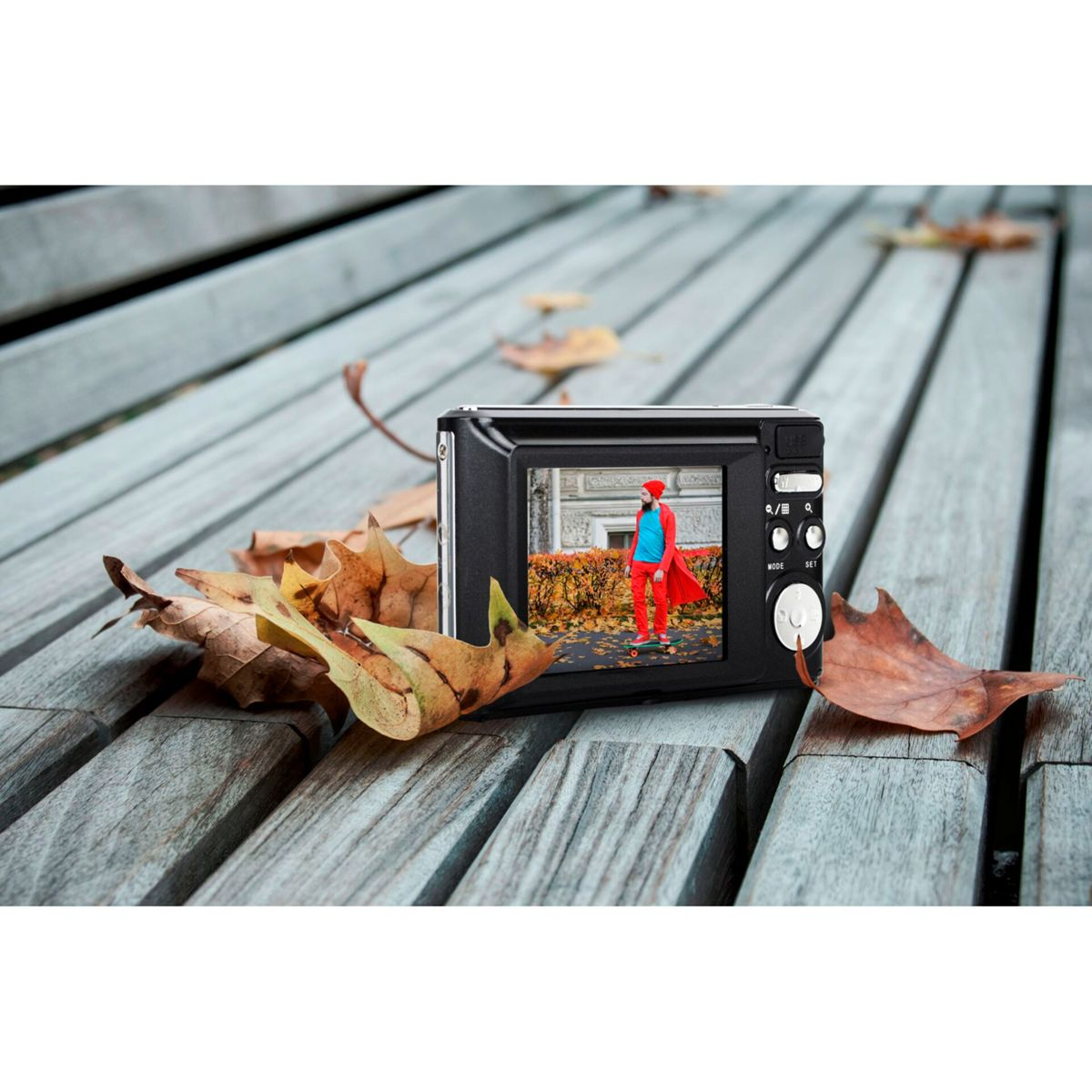 Aparat cyfrowy na drewnianej powierzchni z liśćmi, wyświetlający obraz osoby w czerwonym płaszczu.