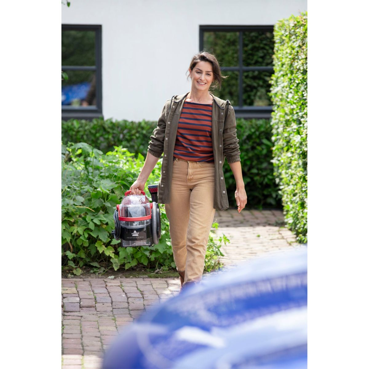 Frau trägt Staubsauger, lächelt, draußen.