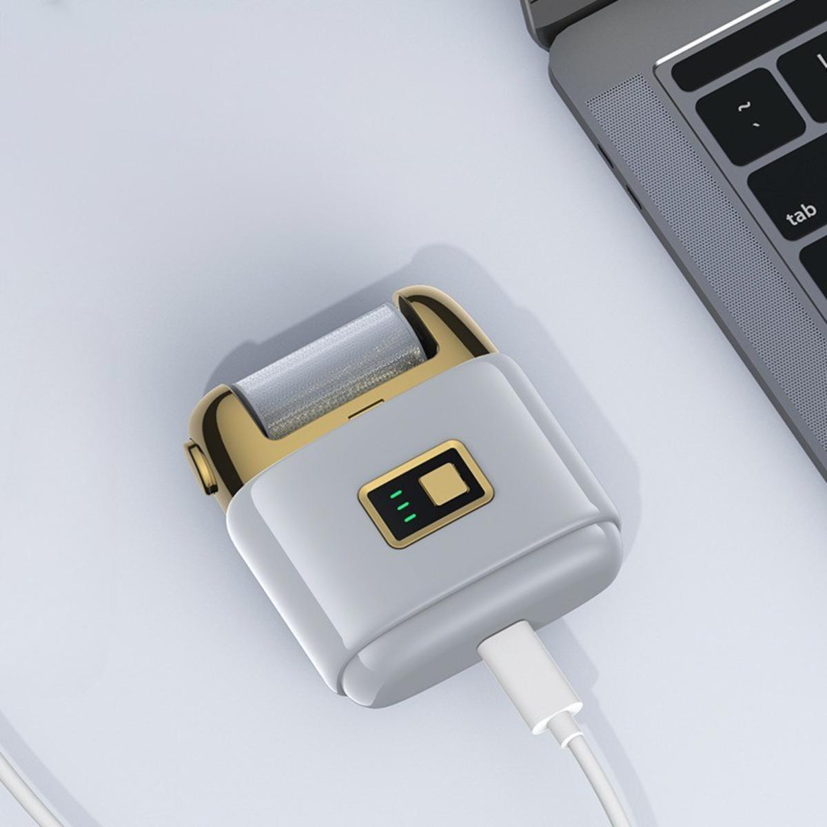 Ein grauer Fußrasierer mit goldenen Akzenten und einem USB-C-Kabel, das an einer hellen Oberfläche in der Nähe eines Laptops liegt.