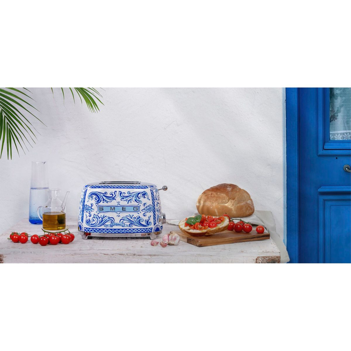 Ein blau-weißer Smeg-Toaster, Brot, Tomaten und Olivenöl stehen auf einem weißen Tisch vor einer weißen Wand.