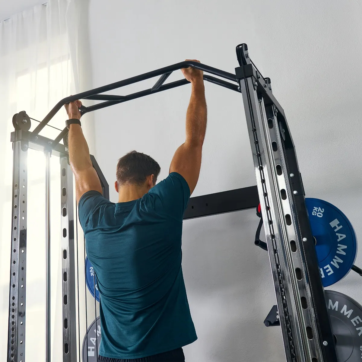 Ein Mann macht Klimmzüge an einer Fitnessmaschine. Er trägt ein blaues Hemd. Der Hintergrund ist eine weiße Wand.