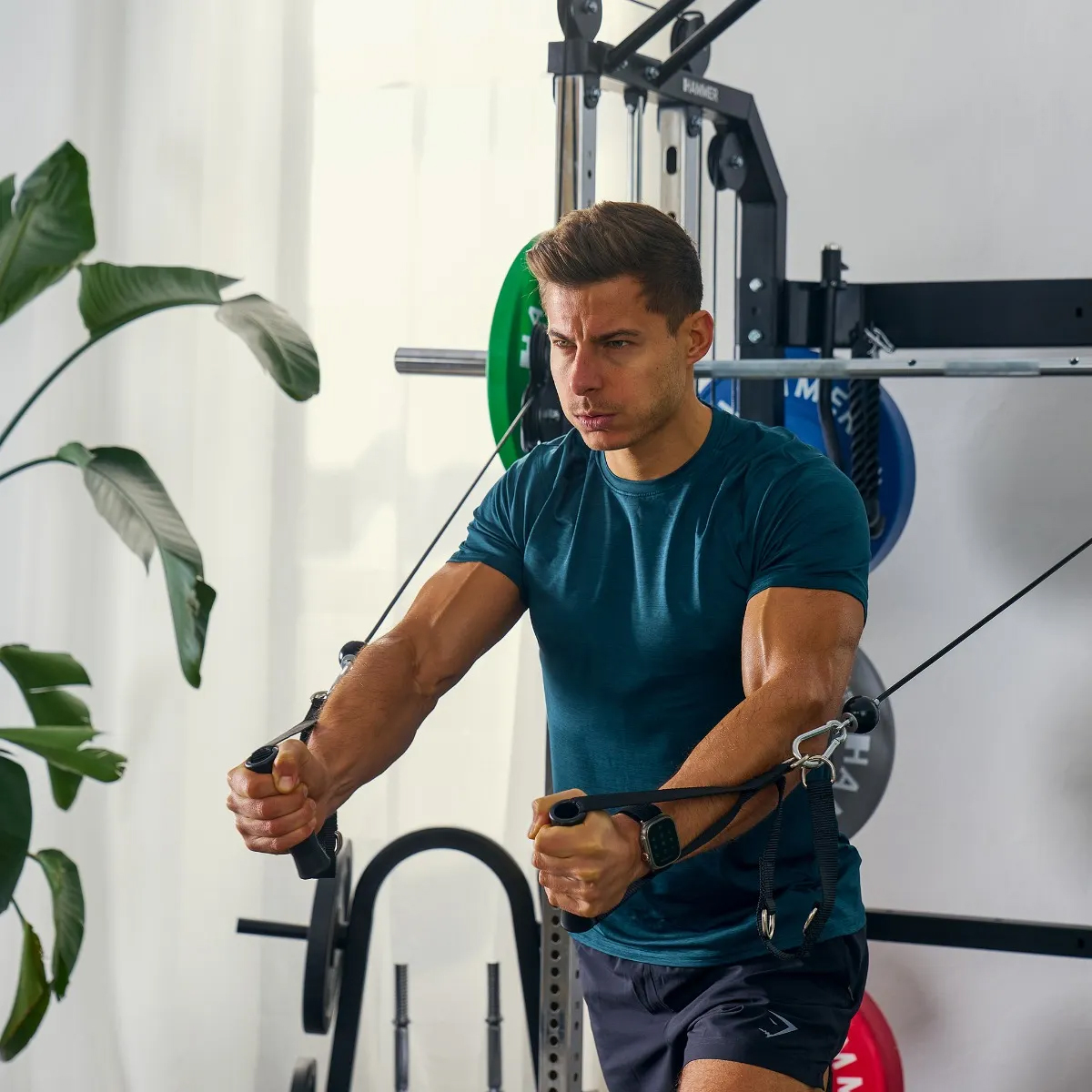 Ein Mann benutzt eine Kabelmaschine in einem Fitnessstudio. Er trägt ein blaues Hemd und schwarze Shorts.