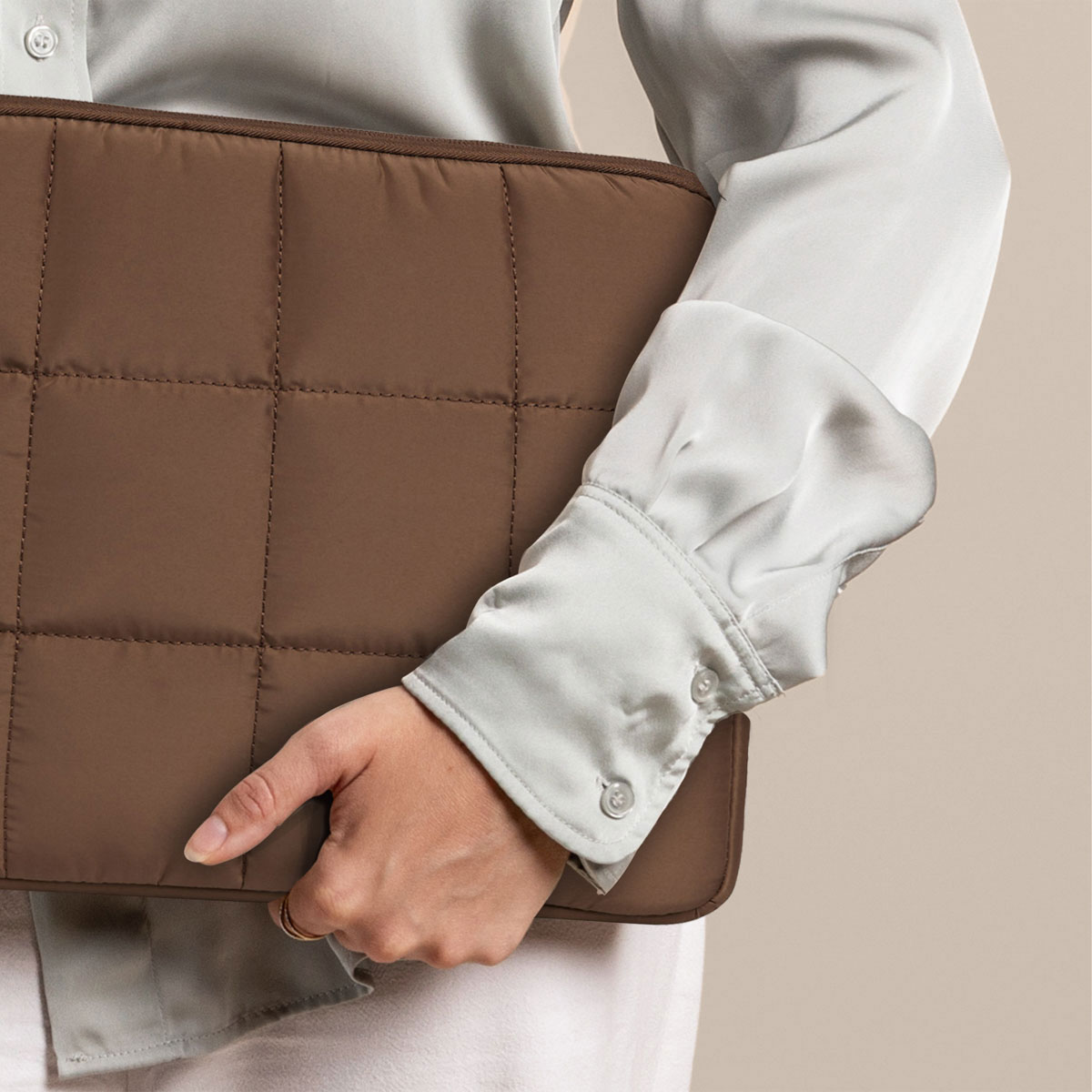 Eine Person hält eine braune, gesteppte Laptop-Hülle. Der Hintergrund ist beige.