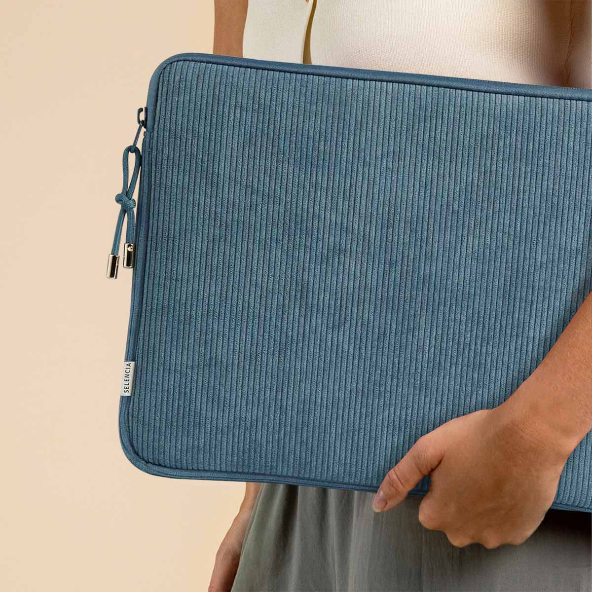 Person hält eine blaue Laptop-Hülle aus Cord. Die Hülle hat einen Reißverschluss und die Marke SELENCIA ist sichtbar.