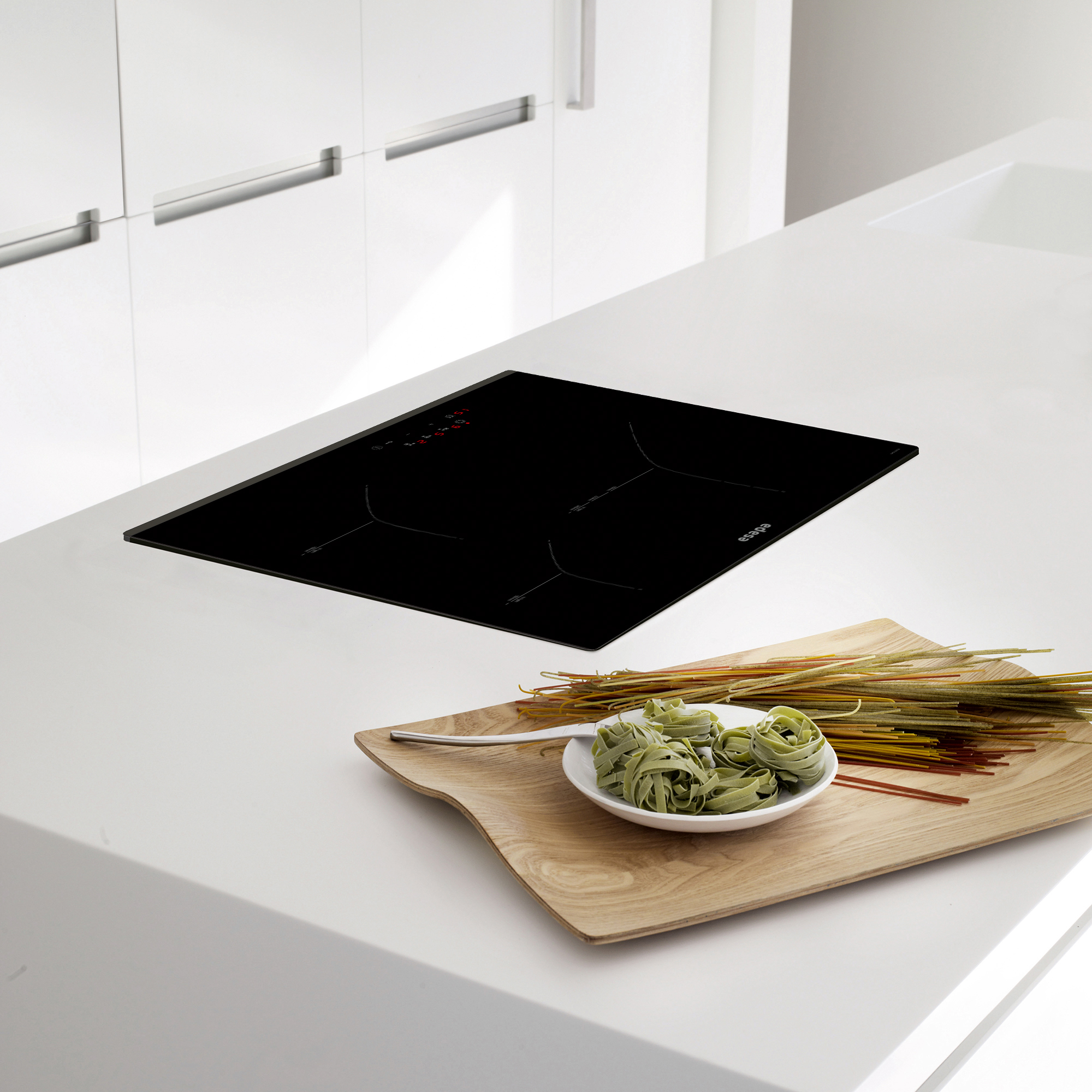 Una escena de cocina moderna con una placa de inducción negra y una bandeja de madera con pasta e ingredientes.