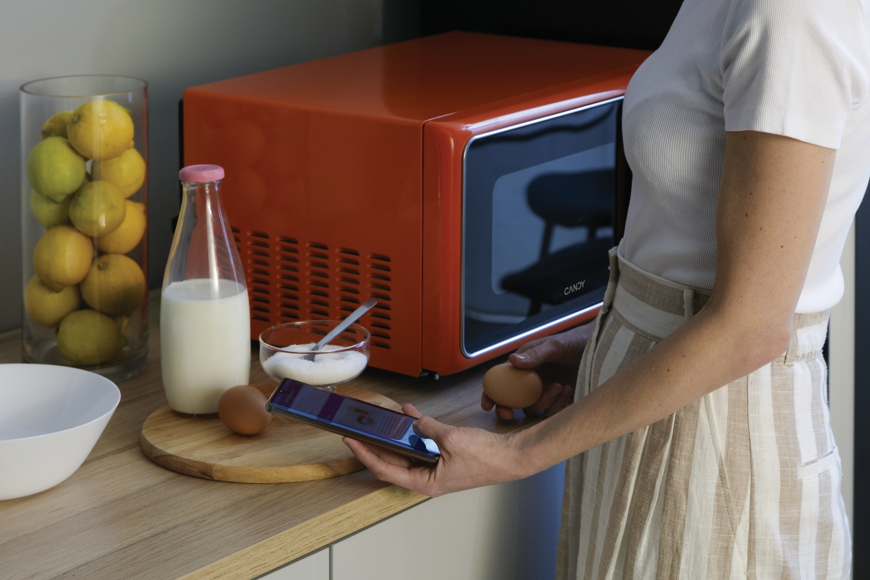 Donna in cucina con microonde arancione. Tiene telefono e uovo. Latte e limoni vicini.