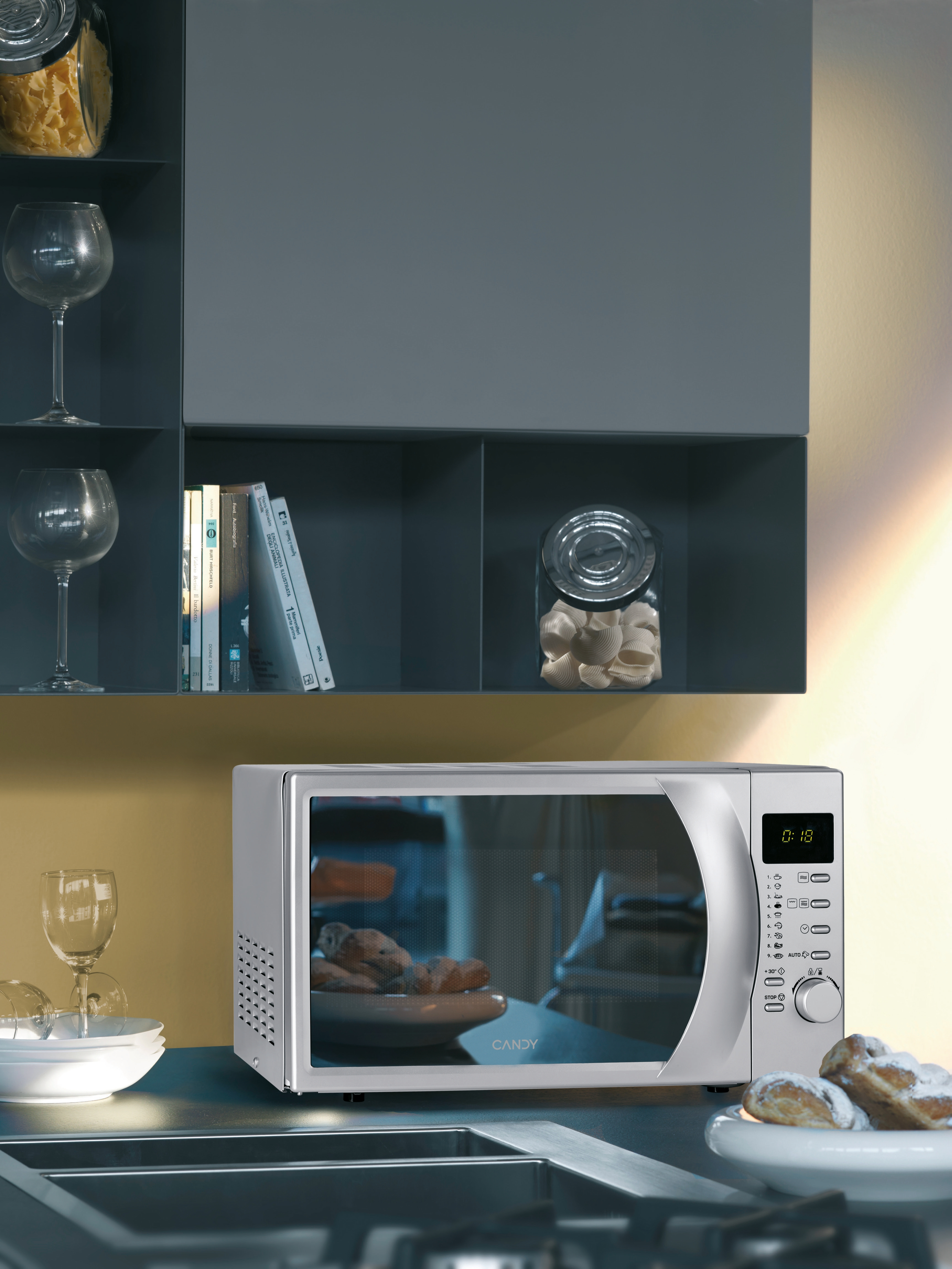 Un forno a microonde su un bancone con un bicchiere di vino e libri sugli scaffali, con un muro giallo sullo sfondo.