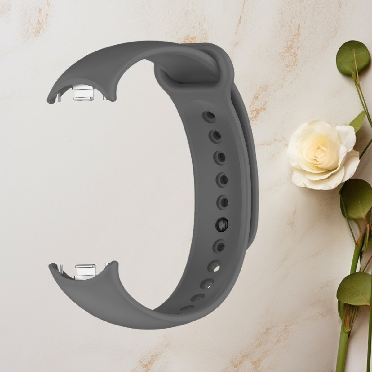 Ein graues Armband mit Verstelllöchern und Metallverschlüssen liegt auf einer beigen Oberfläche mit einer Blume daneben.