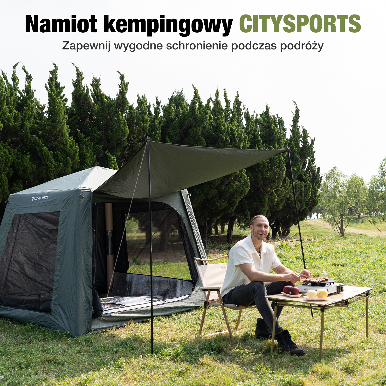 Mężczyzna siedzi przy stole przed zielonym namiotem. Tekst mówi: 'Namiot kempingowy CITYSPORTS'. Drzewa są w tle.