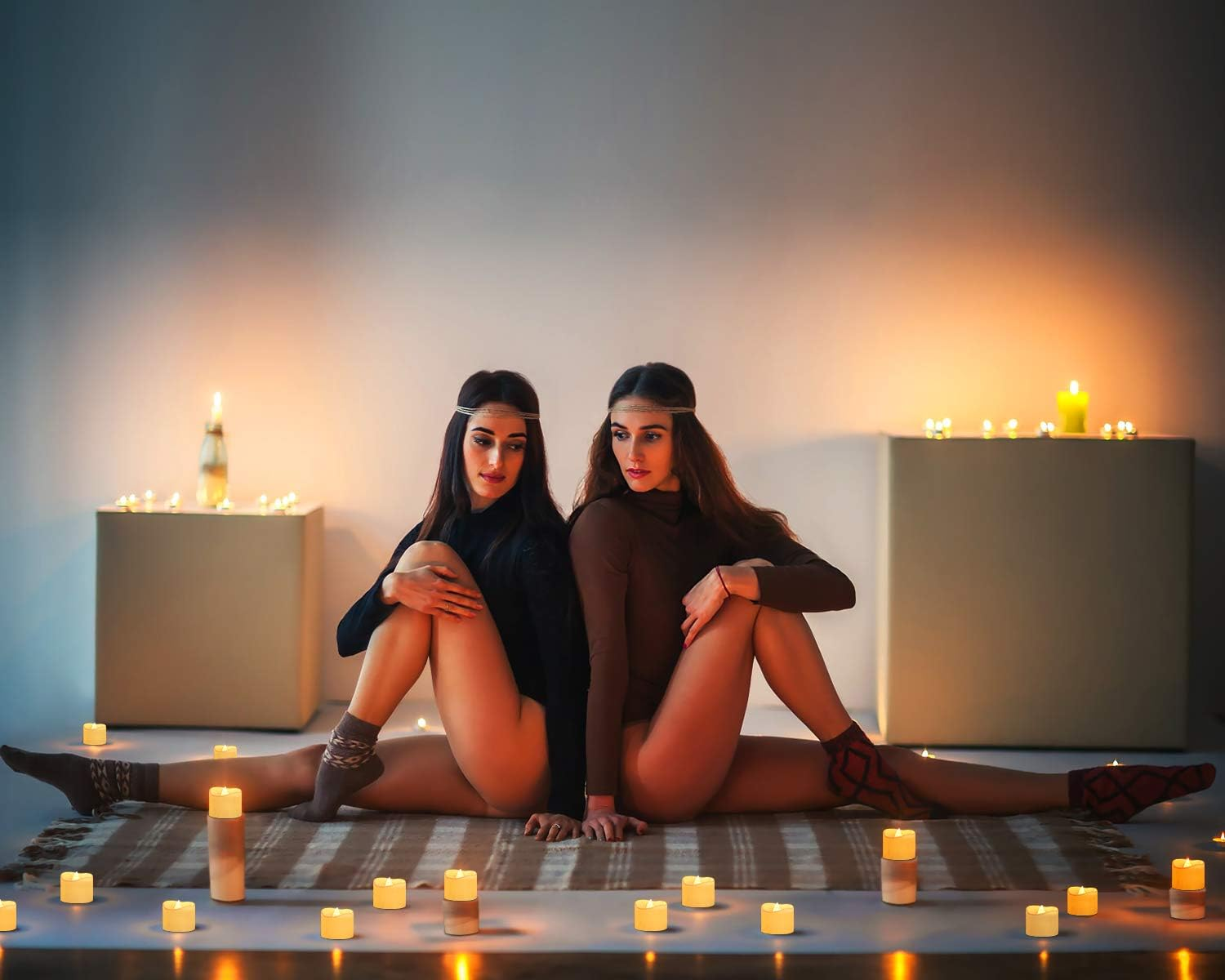 Zwei Frauen in Spagat-Pose, um sie herum Kerzen. Sie tragen Stirnbänder und Socken. Hintergrund: weiße Wand und Boxen.