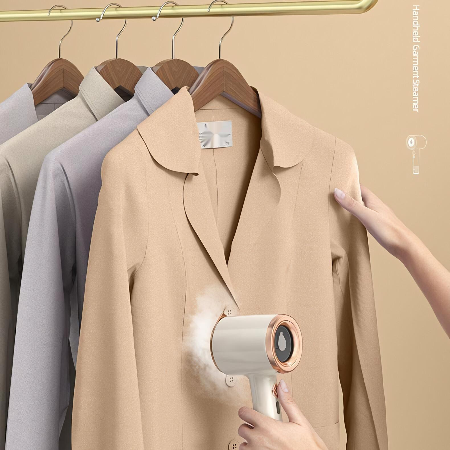 Eine Person dämpft eine beige Jacke mit einem Hand-Dampfgerät. Mäntel hängen an einer goldenen Stange.