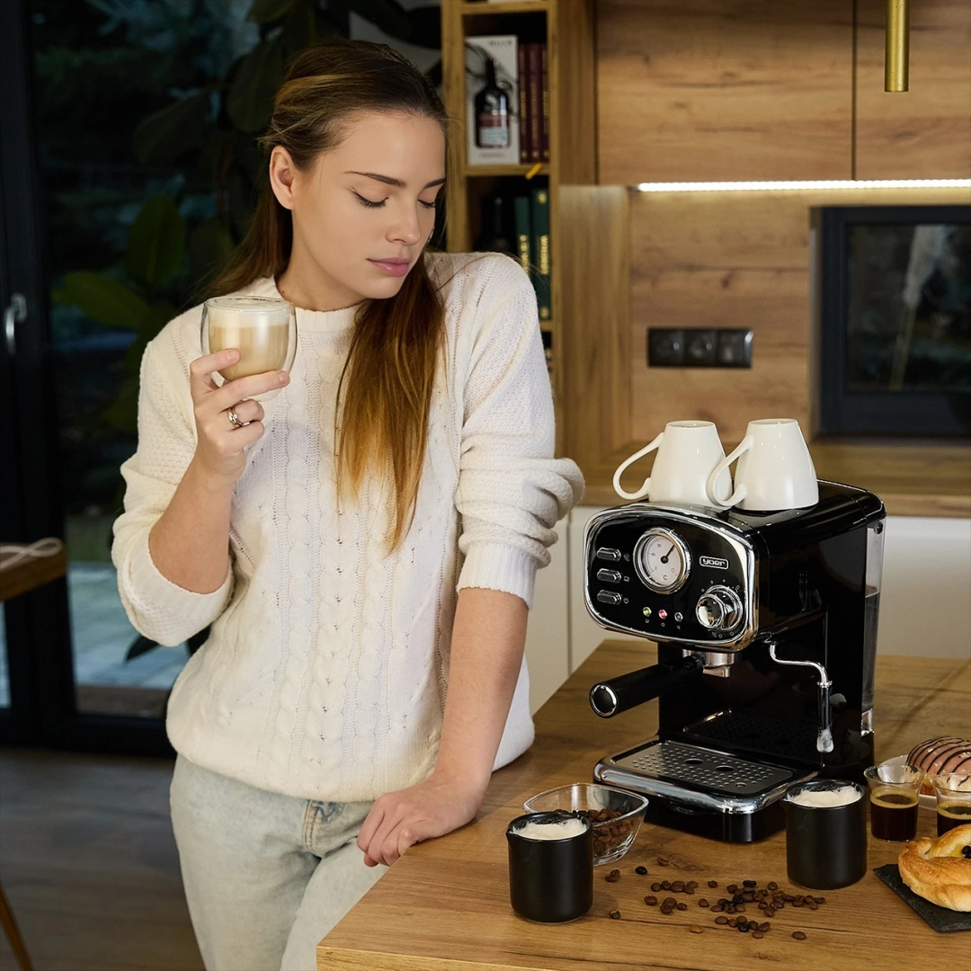 Kobieta stoi przy czarnym ekspresie do kawy, trzymając szklankę kawy. Obecny jest drewniany blat.