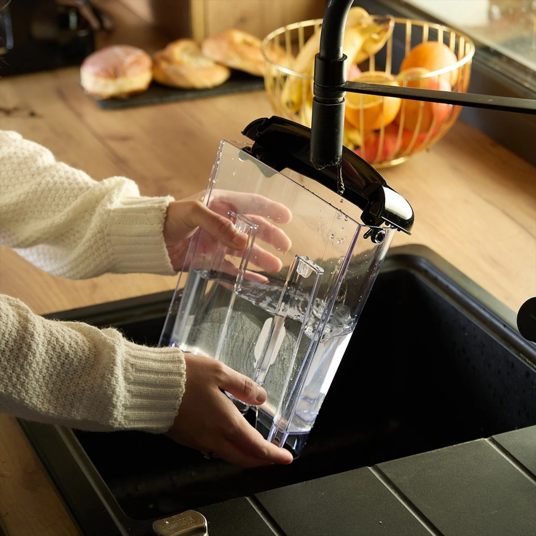 Dłonie trzymające pojemnik na wodę pod kranem w czarnym zlewie.