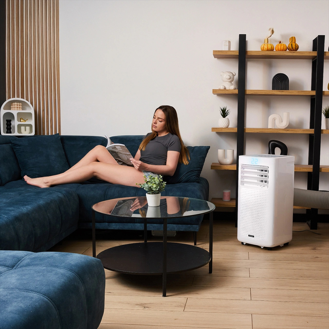 Kobieta czyta na niebieskiej sofie, okrągły stół i biały klimatyzator w salonie.