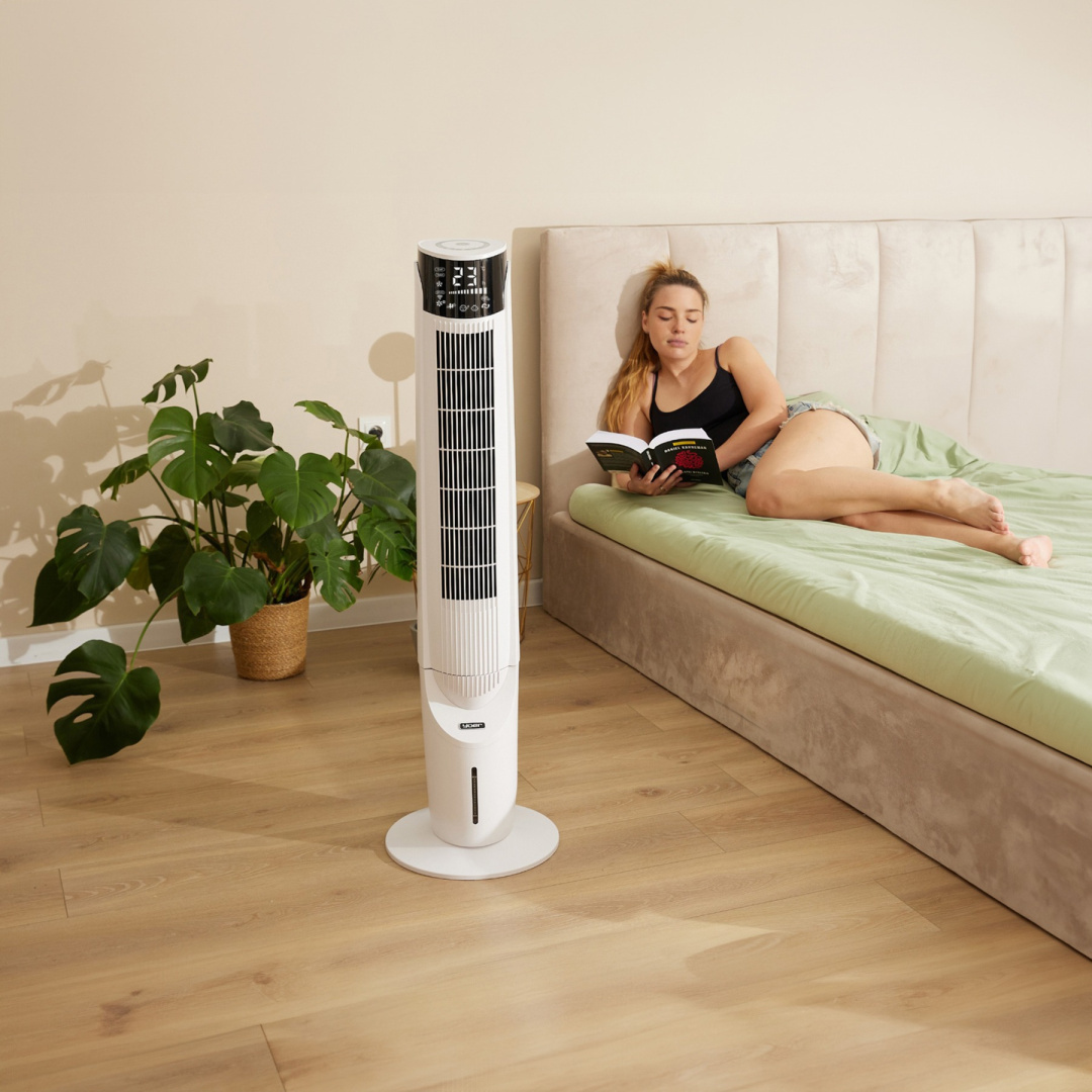 Kobieta czyta na łóżku obok białego wentylatora wieżowego. Roślina na pierwszym planie.