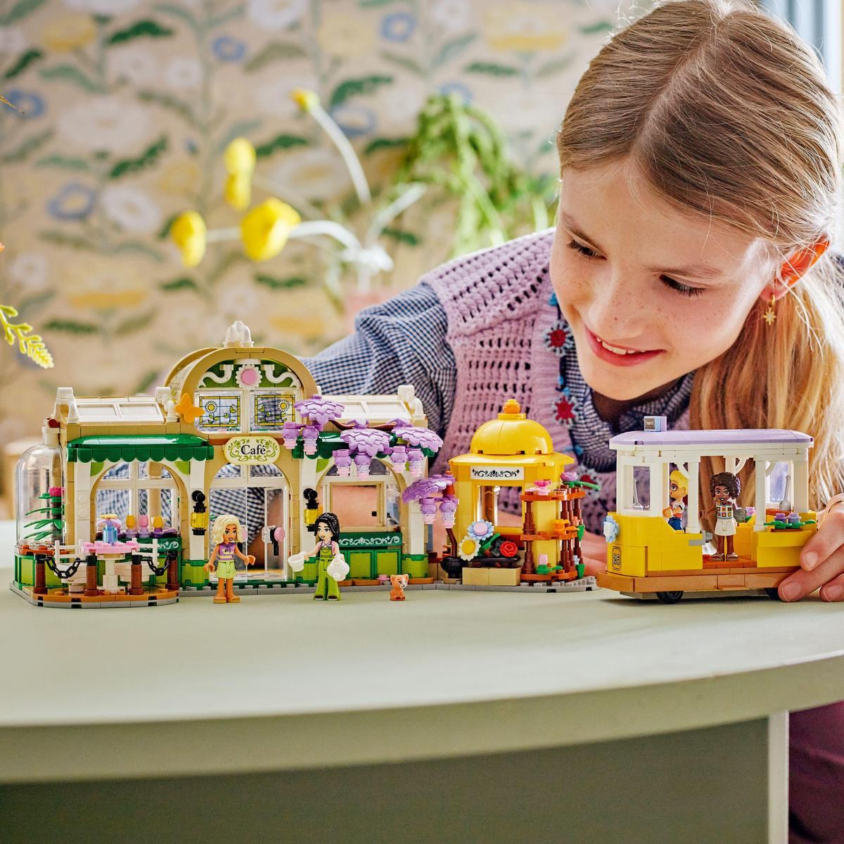 Dziewczyna bawi się zestawem LEGO, z kawiarnią i tramwajem, na tle tapety w kwiaty.