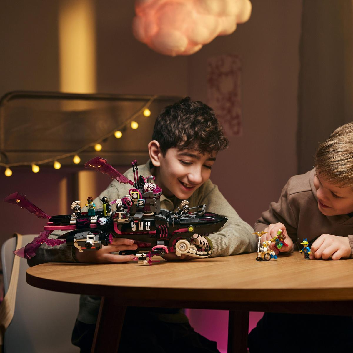 Dwóch chłopców bawiących się czarnym statkiem kosmicznym Lego na drewnianym stole w pokoju.