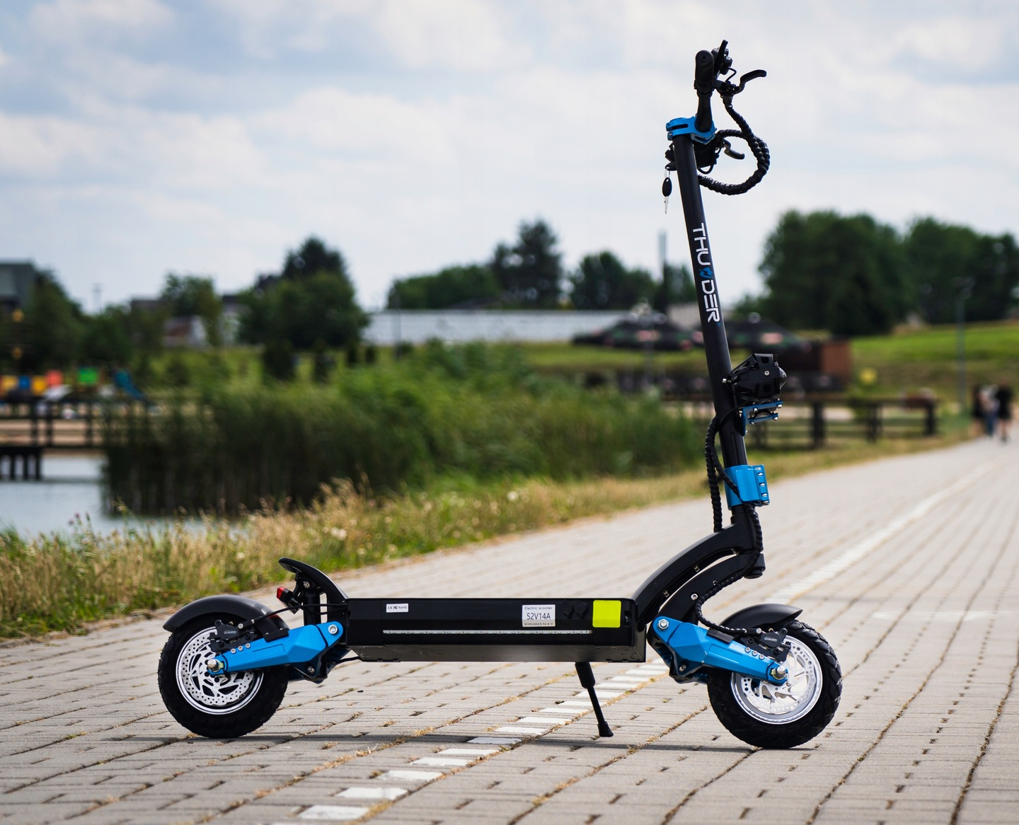 Hulajnoga elektryczna z niebieskimi akcentami zaparkowana na utwardzonej ścieżce, z jeziorem i drzewami w tle.