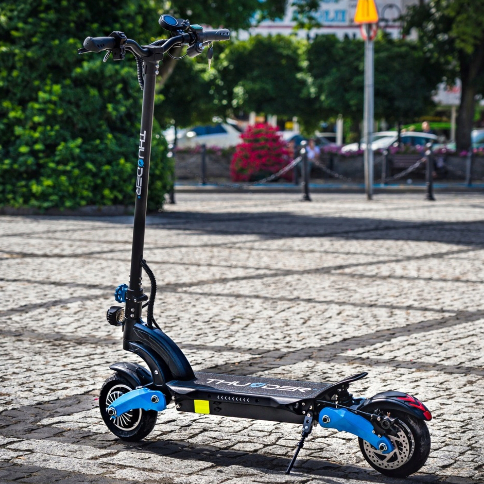Czarno-niebieska hulajnoga elektryczna zaparkowana na utwardzonym terenie, z budynkami i drzewami w tle.
