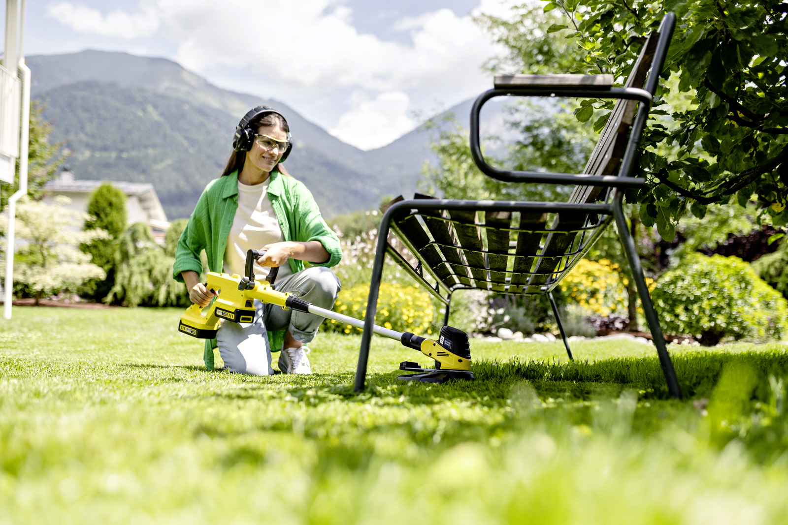 Frau im Garten mit gelbem Rasentrimmer. Sie trägt Schutzbrille und Kopfhörer. Grünes Gras, Berge im Hintergrund.