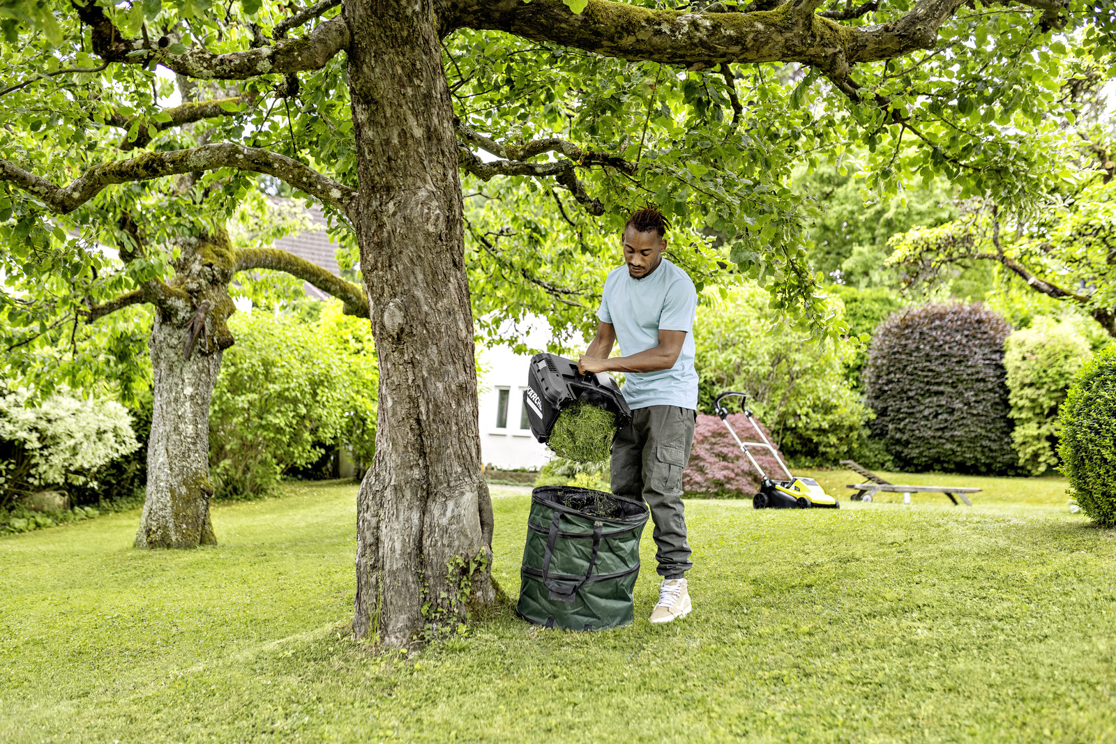 Mann entleert eine grüne Tüte mit Grasschnitt in der Nähe eines Baumes und eines Rasenmähers in einem sonnigen Garten.