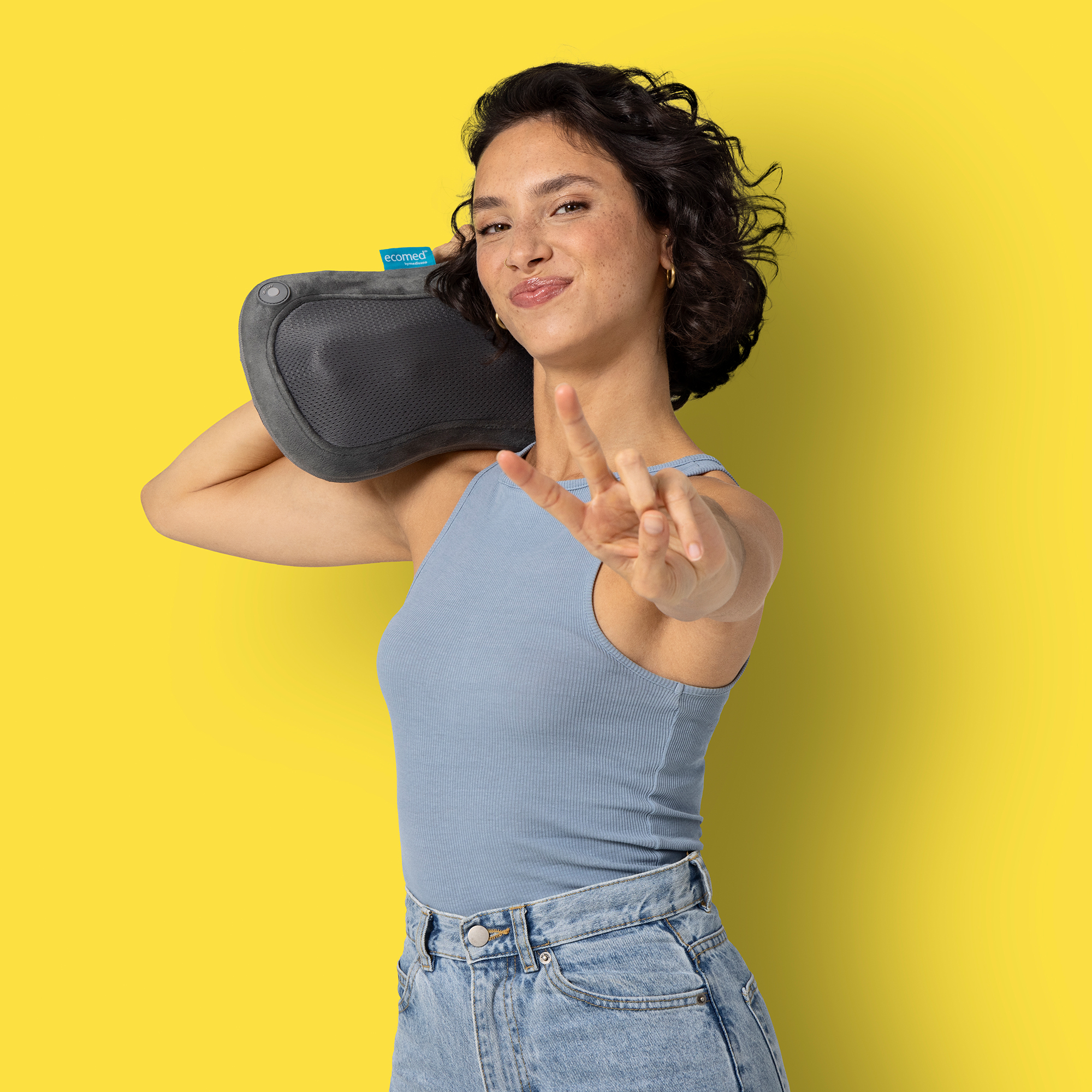 Kobieta trzyma szary masażer szyi i pokazuje znak pokoju na żywym żółtym tle.