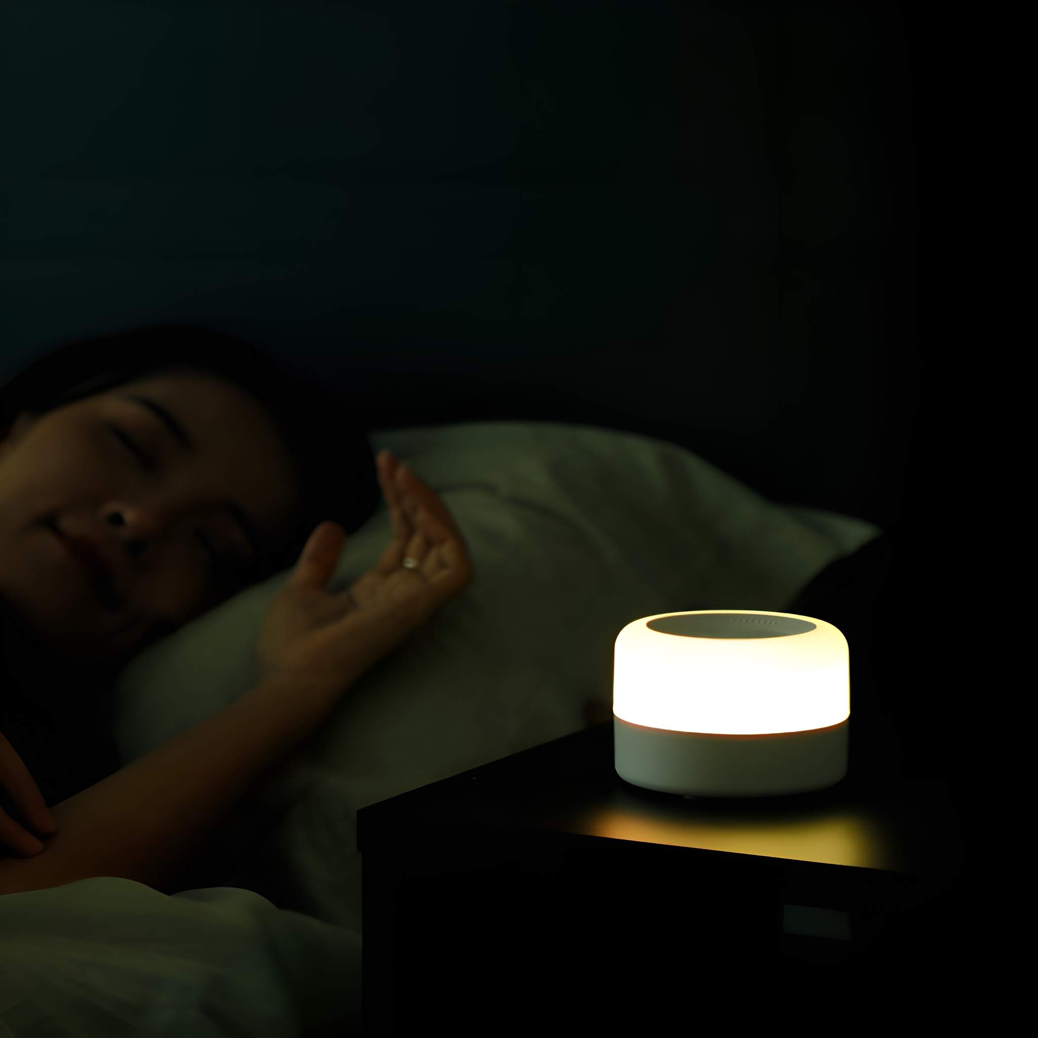 Weiße Lampe auf einem Nachttisch neben einer schlafenden Person. Dunkler Raum, sanftes Leuchten.