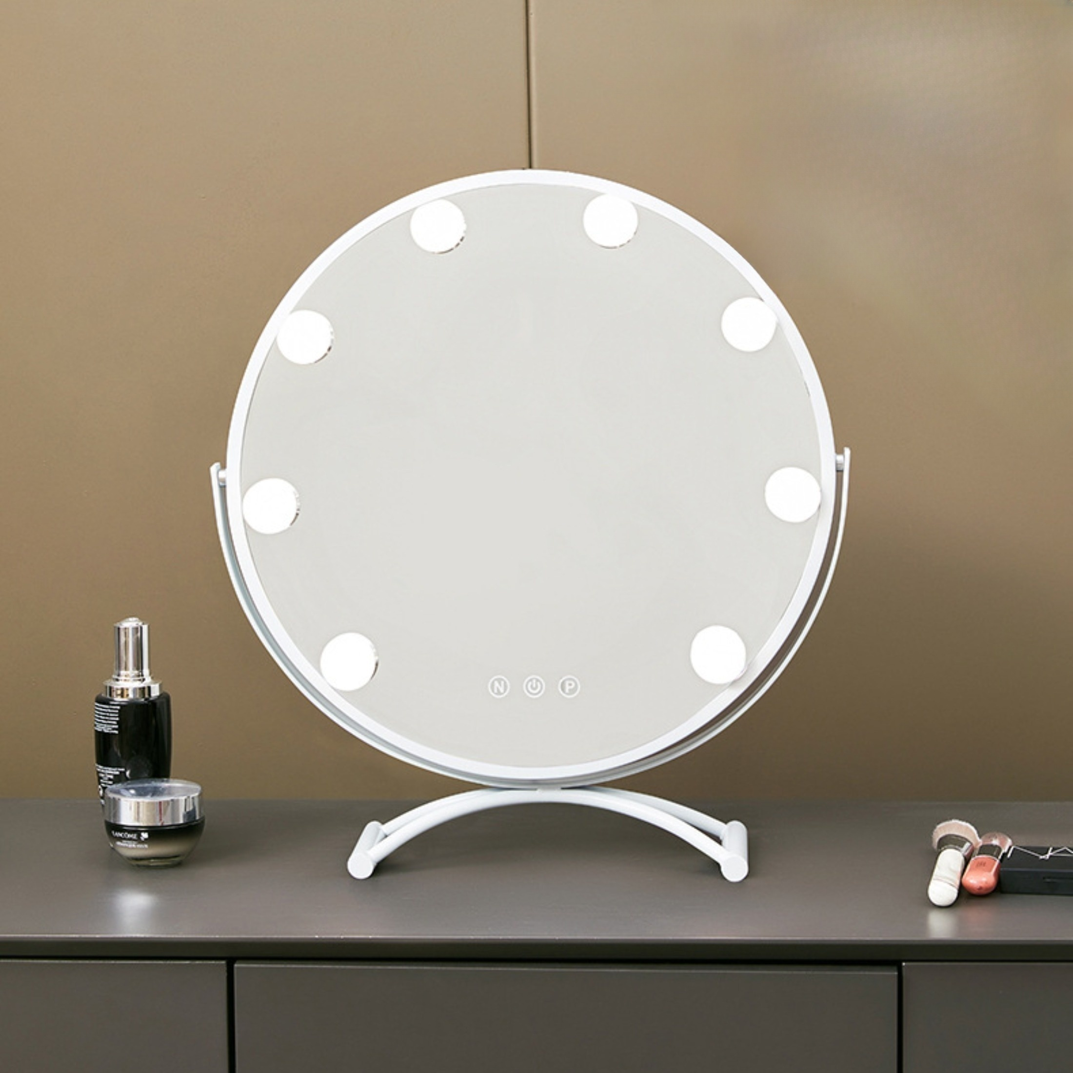 Ein runder Schminkspiegel mit Lichtern steht auf einem Tisch. Im Vordergrund befinden sich Make-up-Artikel.