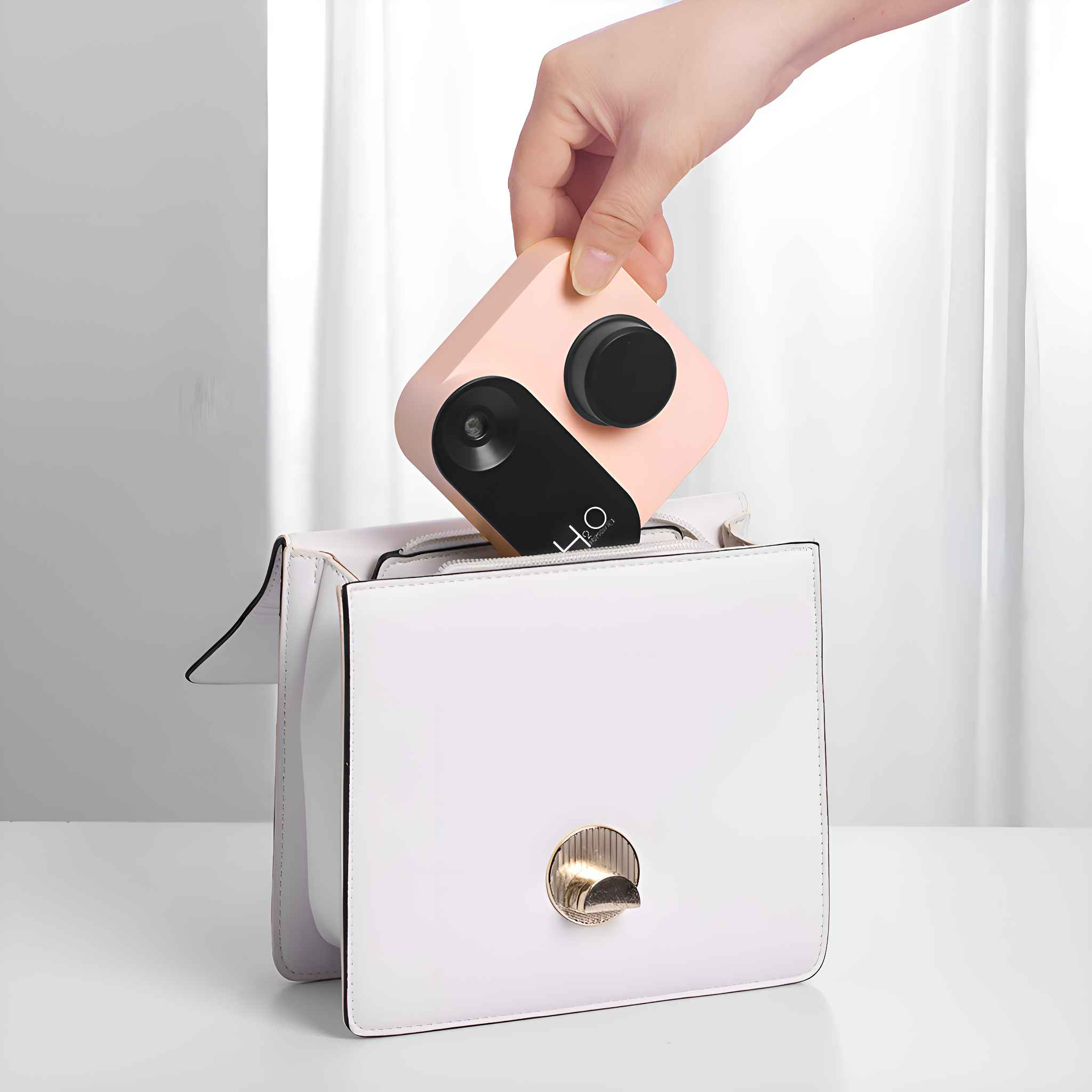 Hand legt ein rosa quadratisches Gerät in eine weiße Handtasche. Schwarzer Kreis und Quadrat oben.