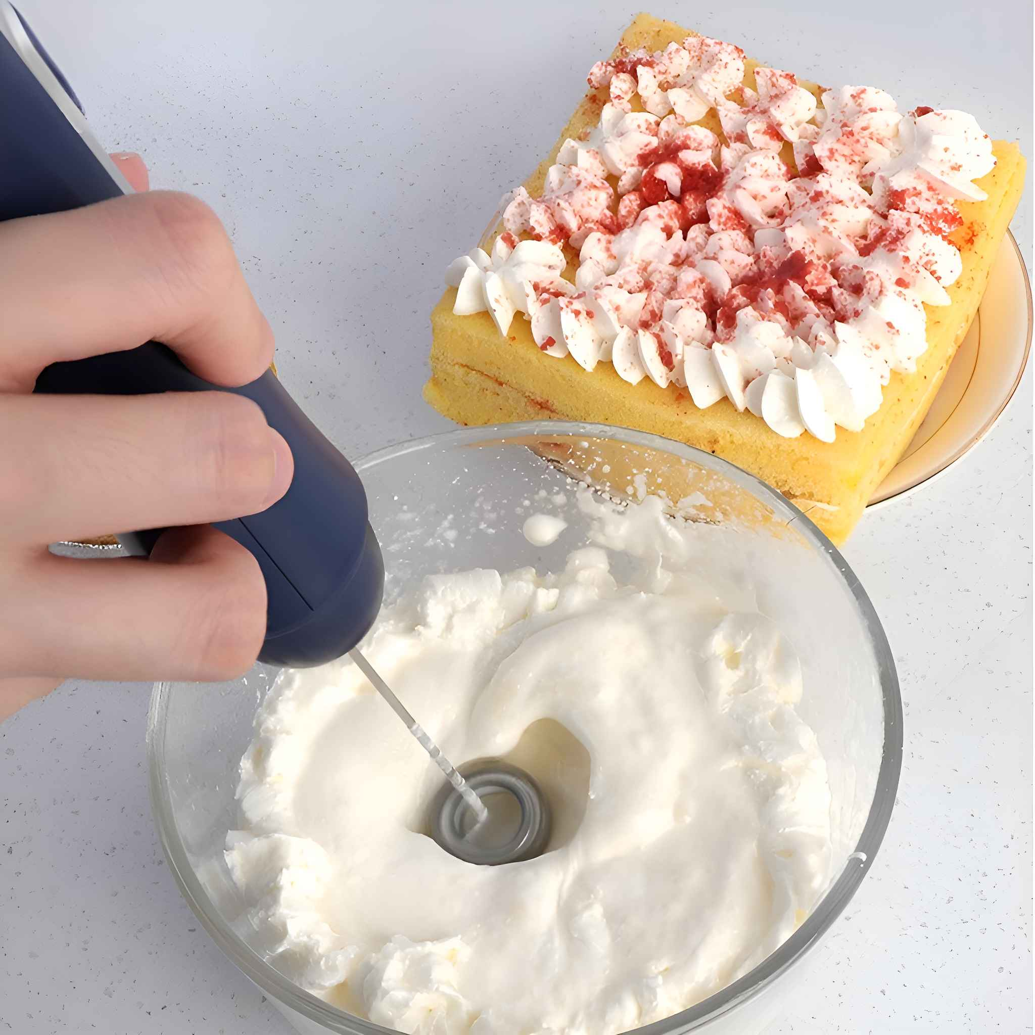 Eine Hand benutzt einen Aufschäumer, um weiße Sahne in einer Schüssel zu verrühren, rechts ein Kuchenstück mit Schlagsahne.