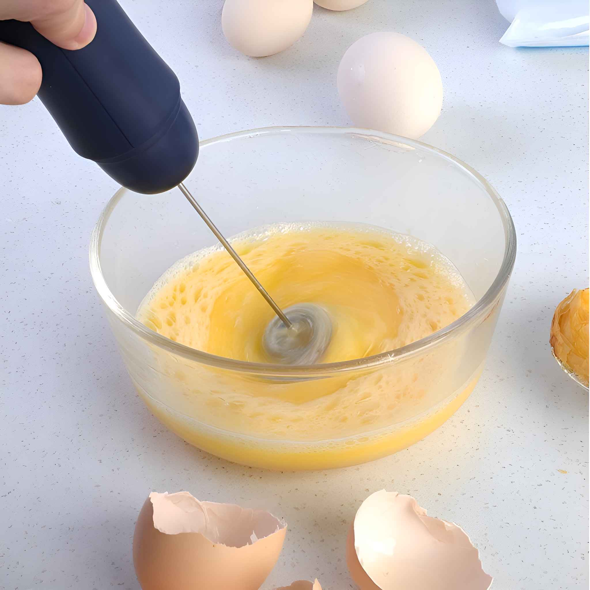 Eine Hand verwendet einen Aufschäumer, um Eier in einer Glasschüssel zu verrühren. Einige Eier liegen auf dem Tisch, eine Eierschale ist sichtbar.
