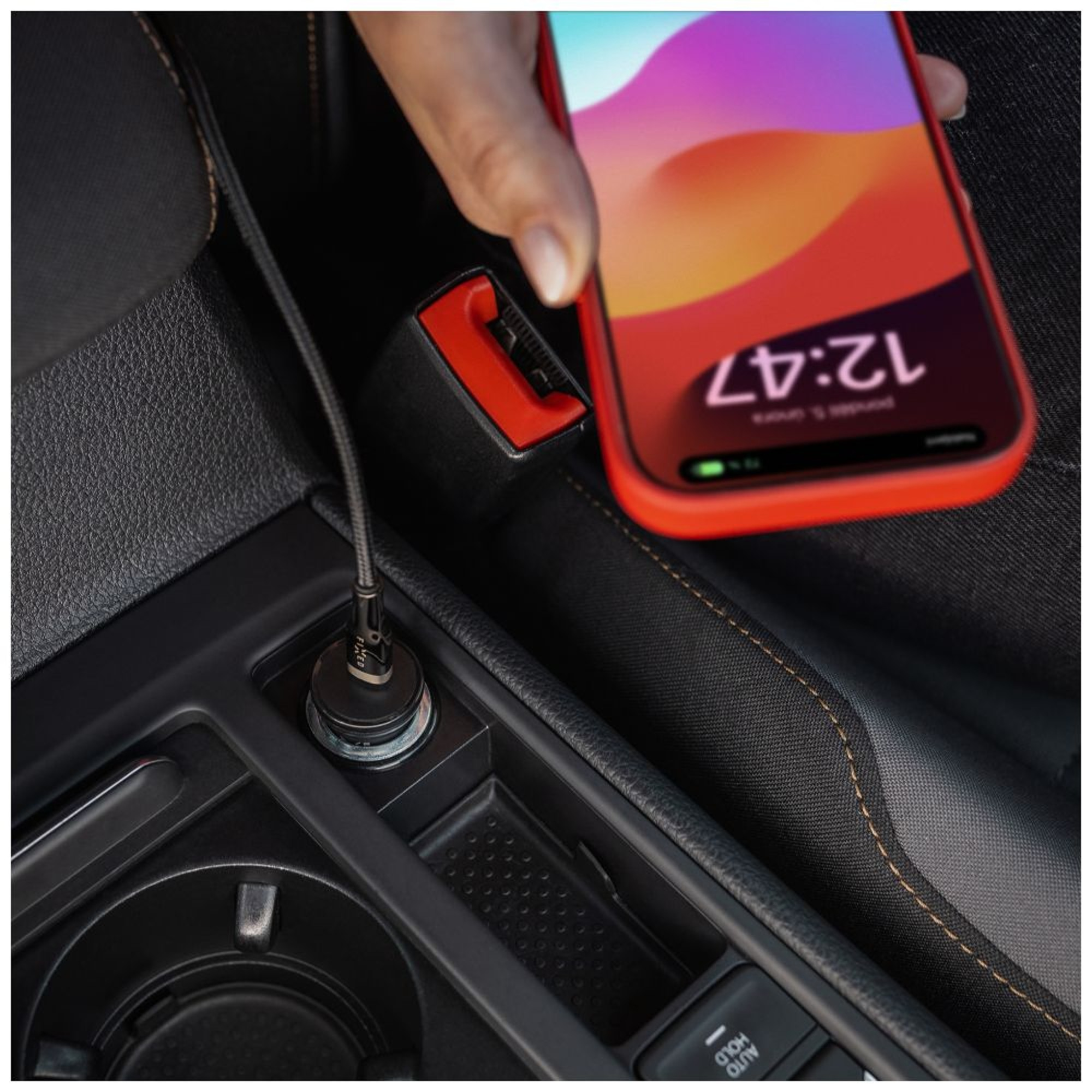 Hand, die ein rotes Smartphone hält, das an ein Autoladegerät angeschlossen ist. Das Telefon zeigt 12:47 Uhr.