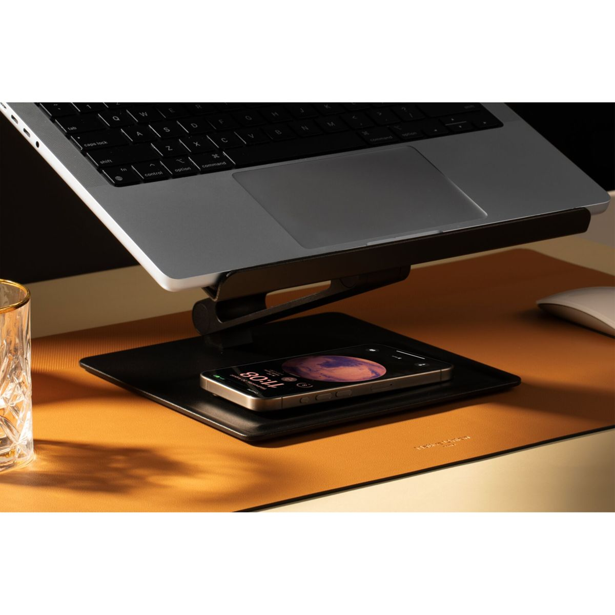 Un laptop su un supporto con un telefono su un vassoio, vicino a un bicchiere e un mouse.