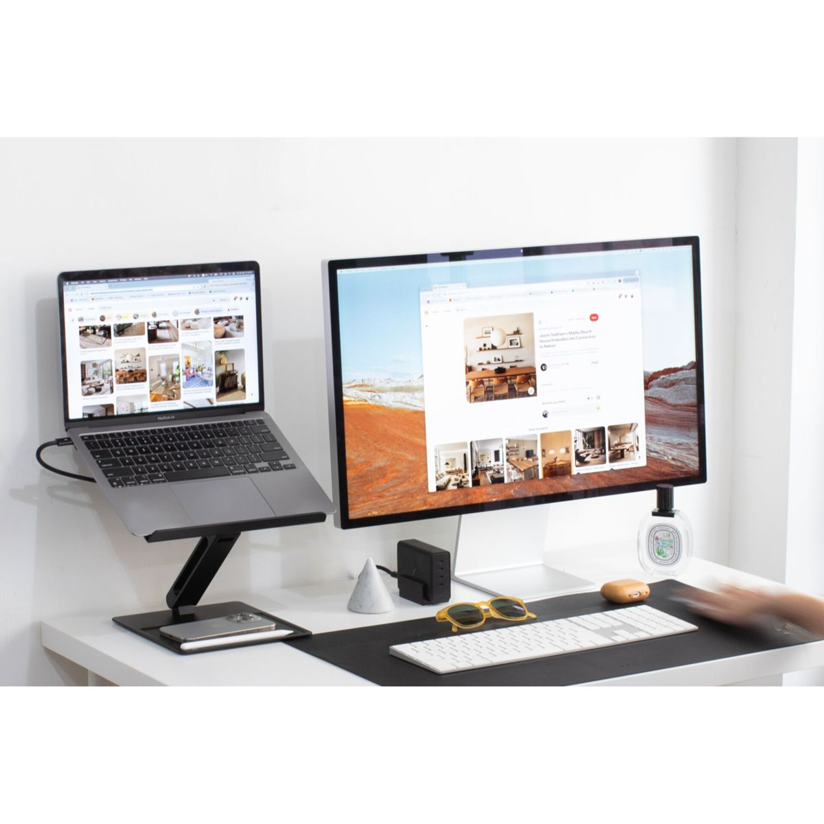 Un laptop su un supporto accanto a un monitor. Una scrivania con accessori, un muro bianco sullo sfondo.