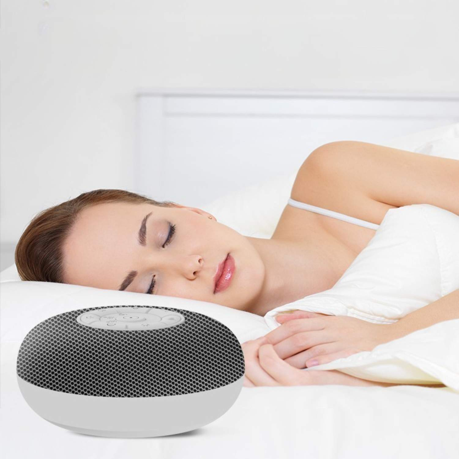 Eine Frau schläft im Bett, im Vordergrund eine Geräuschmaschine. Die Maschine ist grau und rund.