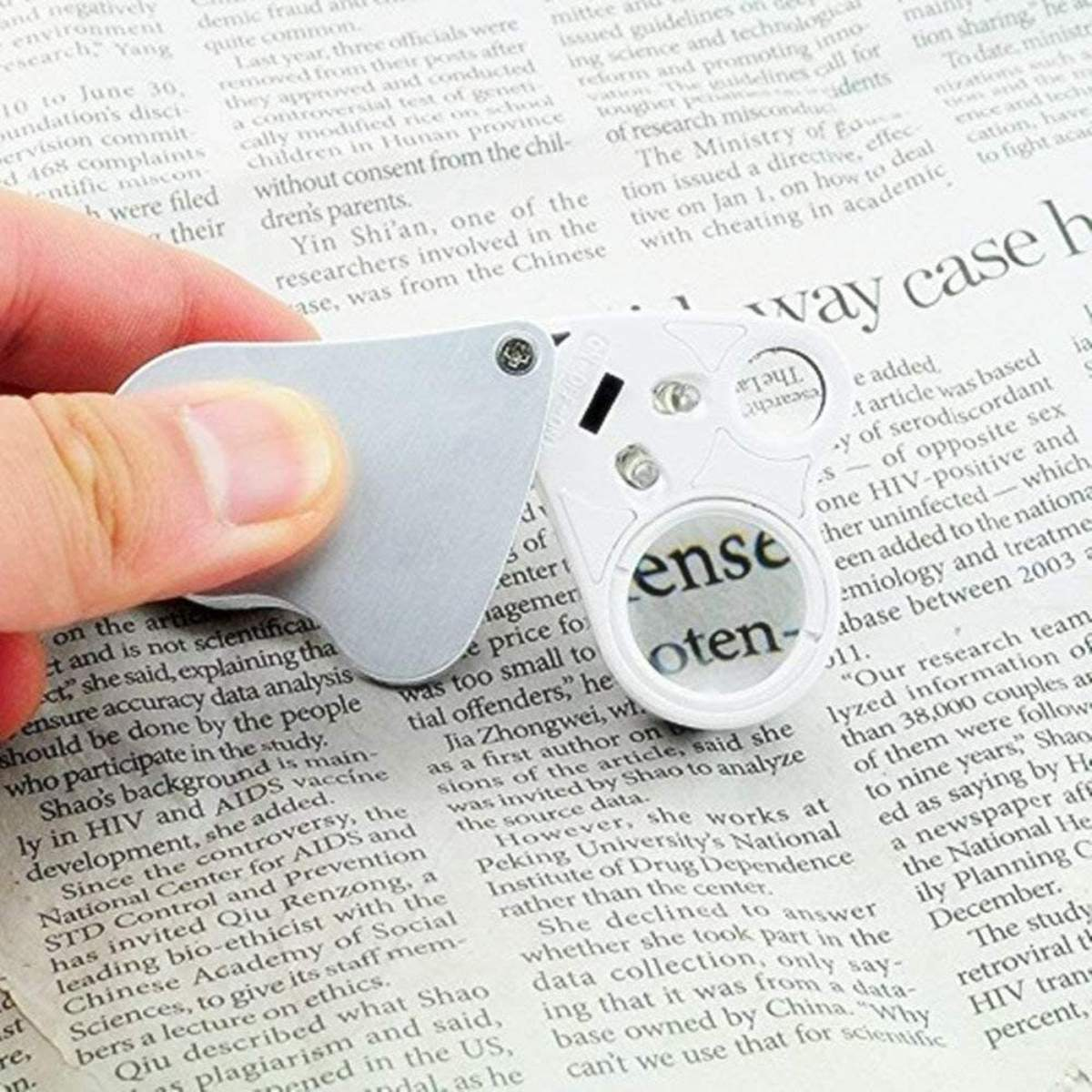 Hand hält eine Lupe über einer Zeitung. Die Lupe ist weiß und silber, mit einer kreisförmigen Linse.