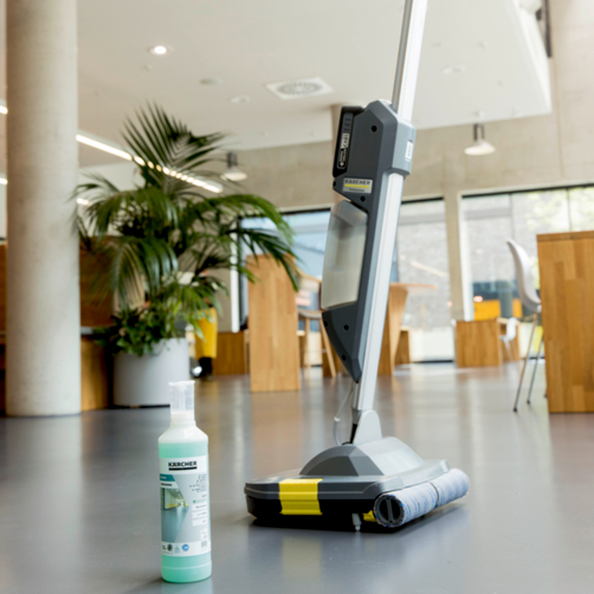 Myjka do podłóg stoi z butelką roztworu czyszczącego. W pokoju jest duża roślina i okno.