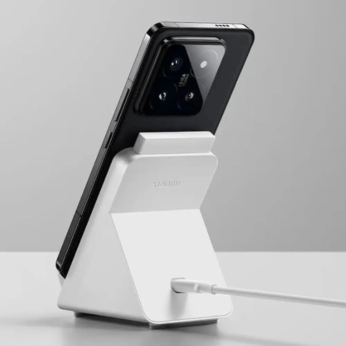 Black smartphone on a white charging stand with a cable. Leica camera visible. The background is a light gray.