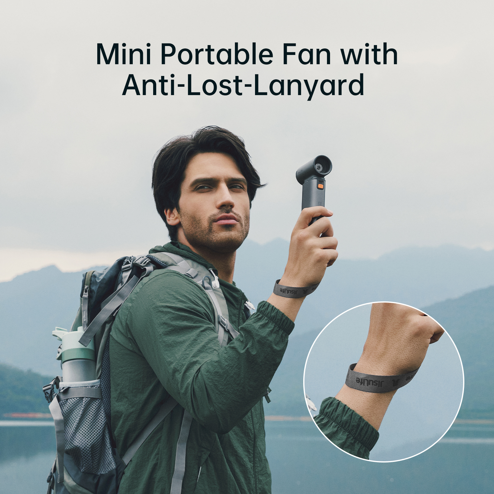 Hombre sosteniendo un mini ventilador. Lleva una chaqueta verde, una mochila. Montañas y un lago al fondo.