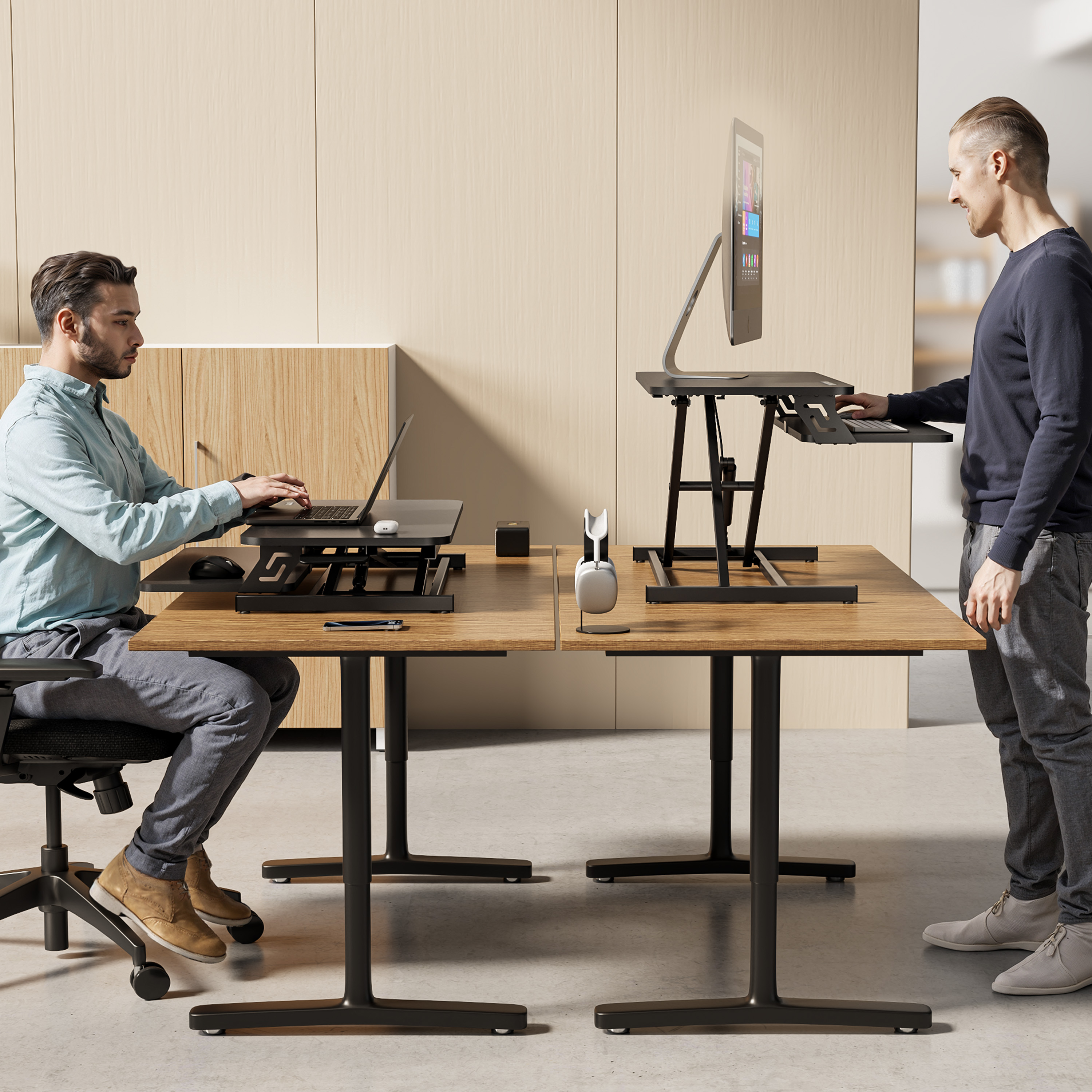 Zwei Männer an Schreibtischen. Einer sitzt und benutzt einen Laptop, der andere steht mit einem Computer. Neutraler Hintergrund.