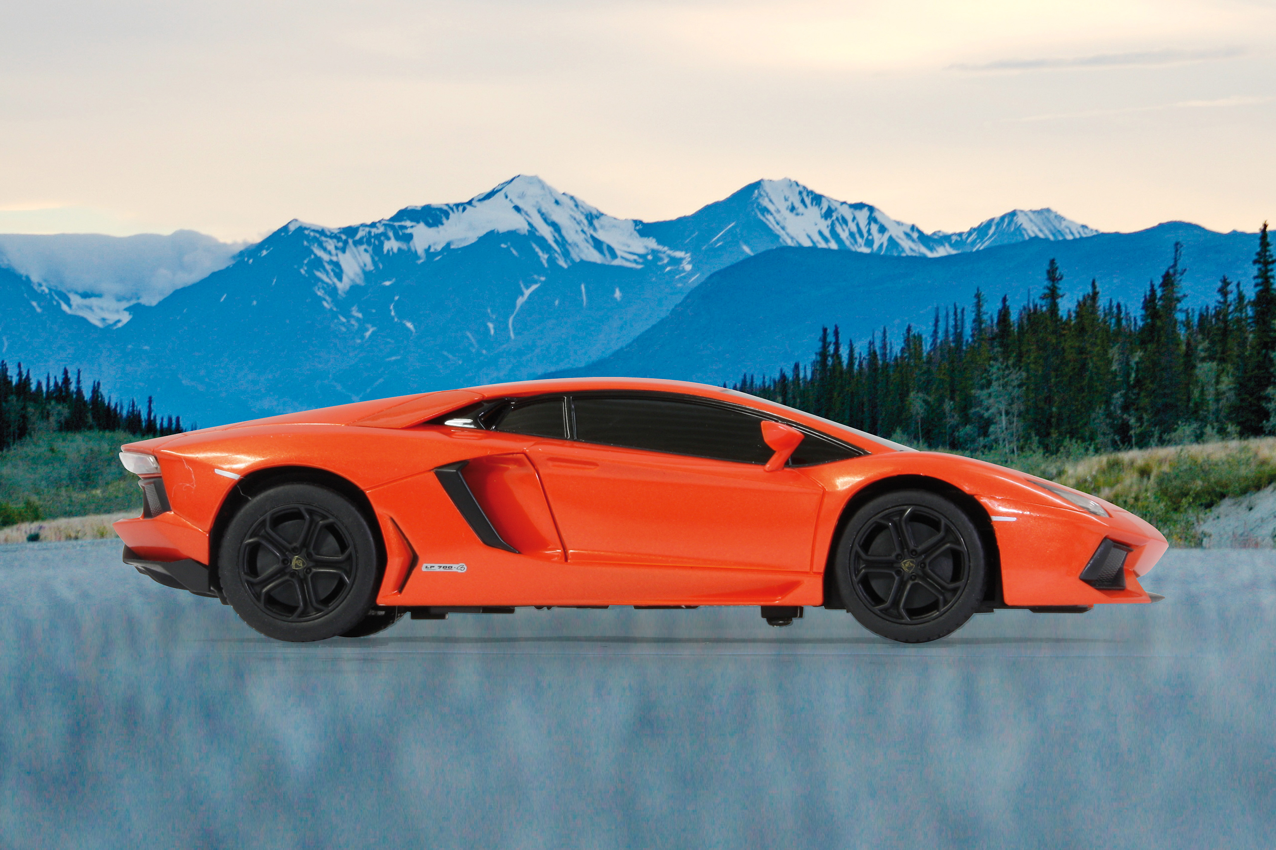 Oranger Lamborghini-Spielzeugwagen auf einer hellblauen, reflektierenden Oberfläche, mit Berg- und Himmelshintergrund. Schwarze Räder.