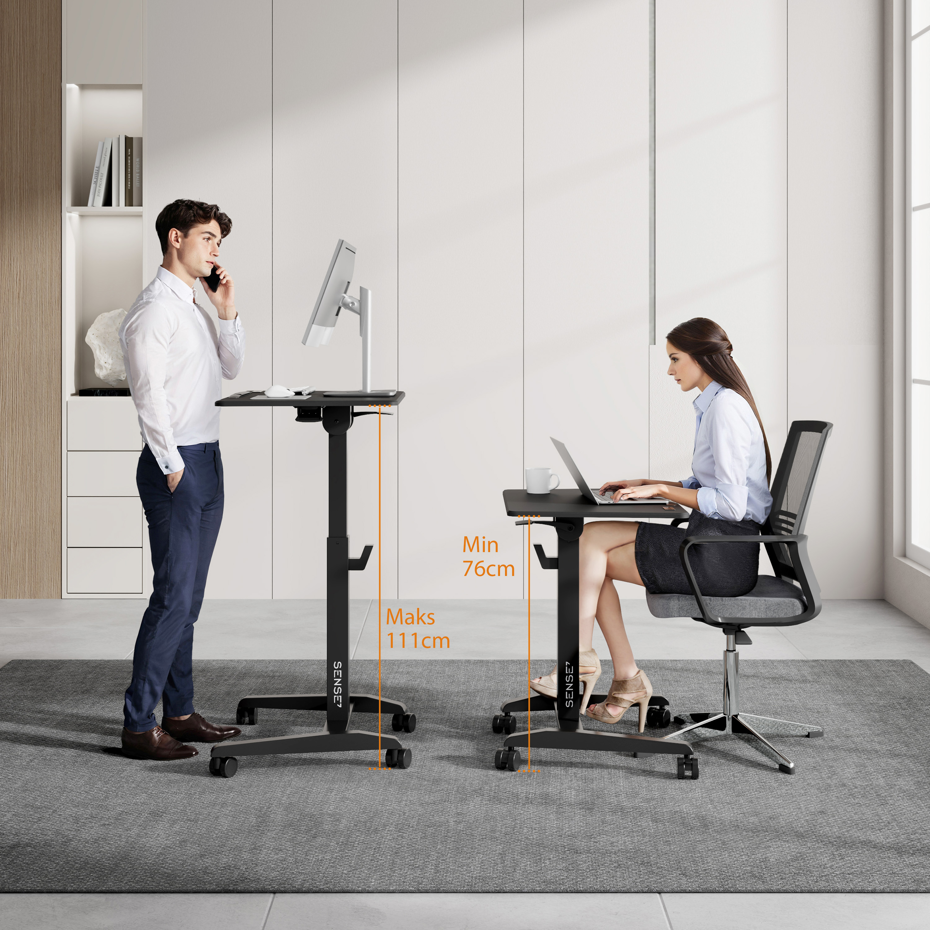 Zwei Personen an höhenverstellbaren Schreibtischen. Einer telefoniert, der andere arbeitet am Laptop. Grauer Teppichboden.