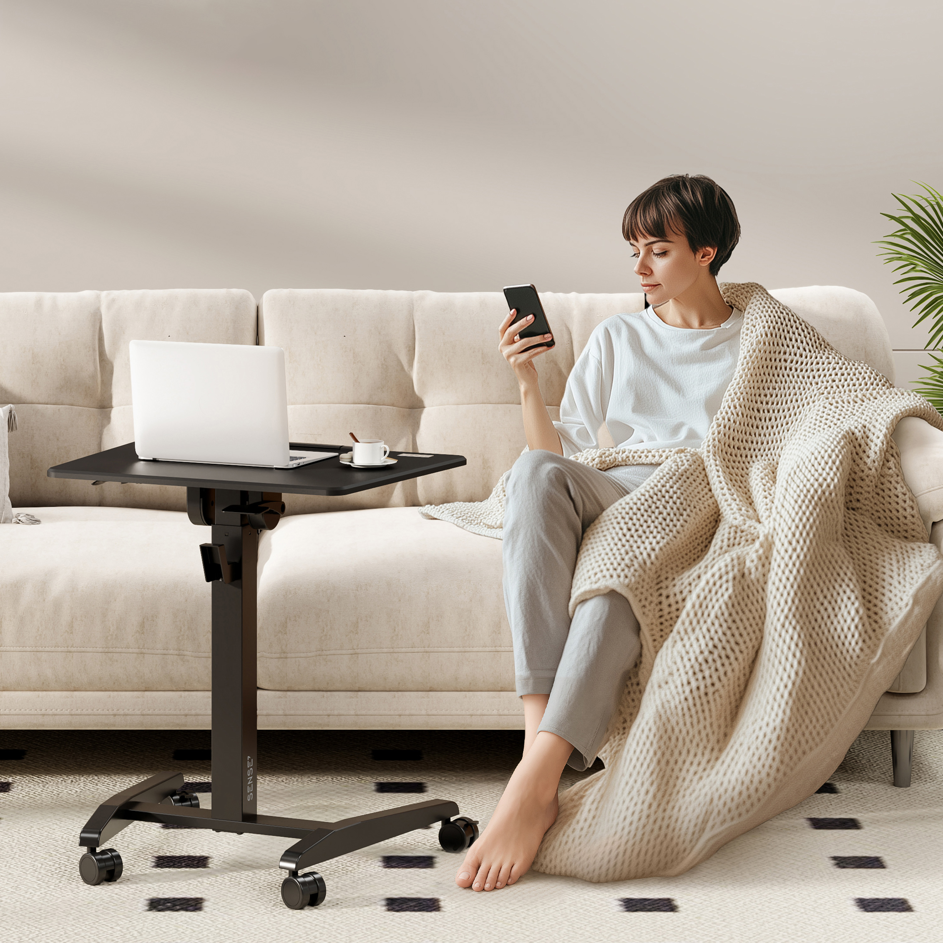 Mujer en sofá con teléfono, portátil en mesa. Manta la cubre. La habitación tiene una alfombra y una planta.